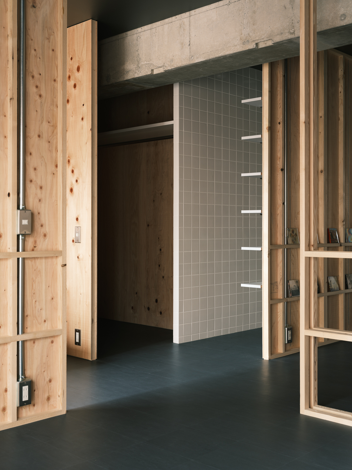 Open timber door revealing a hallway with grey tiled wall and exposed concrete beam overhead