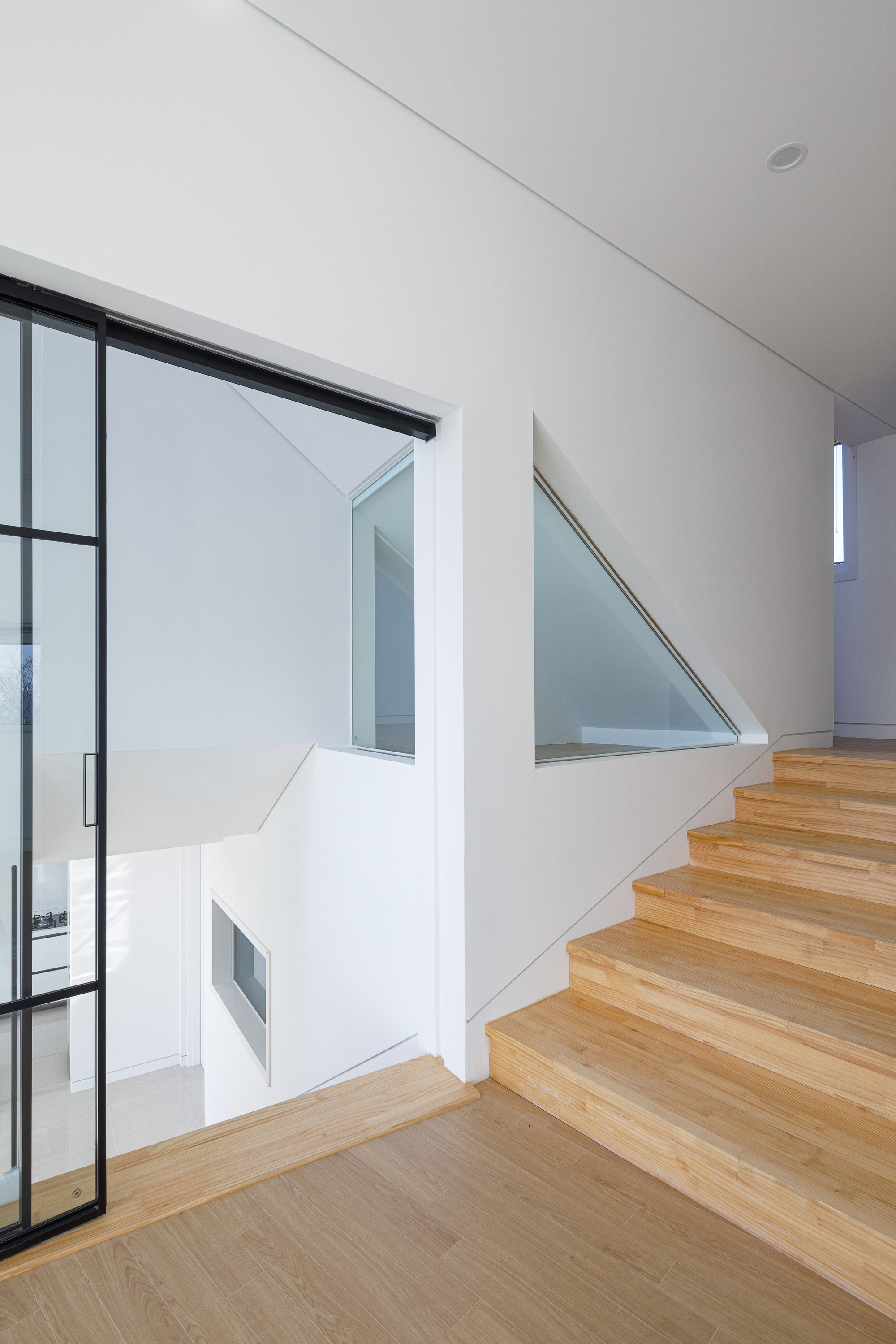 Timber staircase ascending between white walls with steel-framed glass partitions and triangular clerestory windows