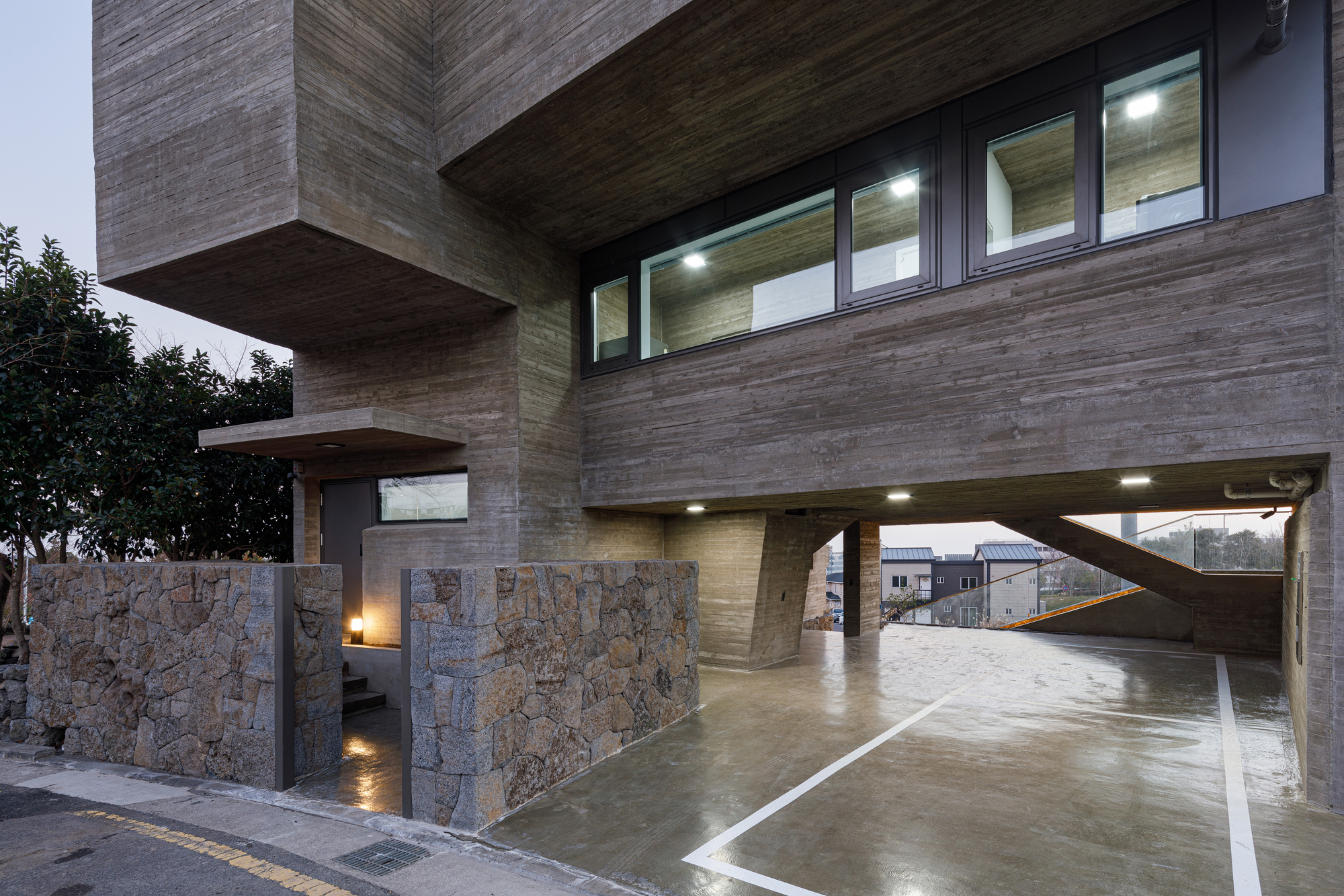 Horizontal timber-clad facade on pilotis above stone-clad retaining walls and wet concrete driveway at dusk
