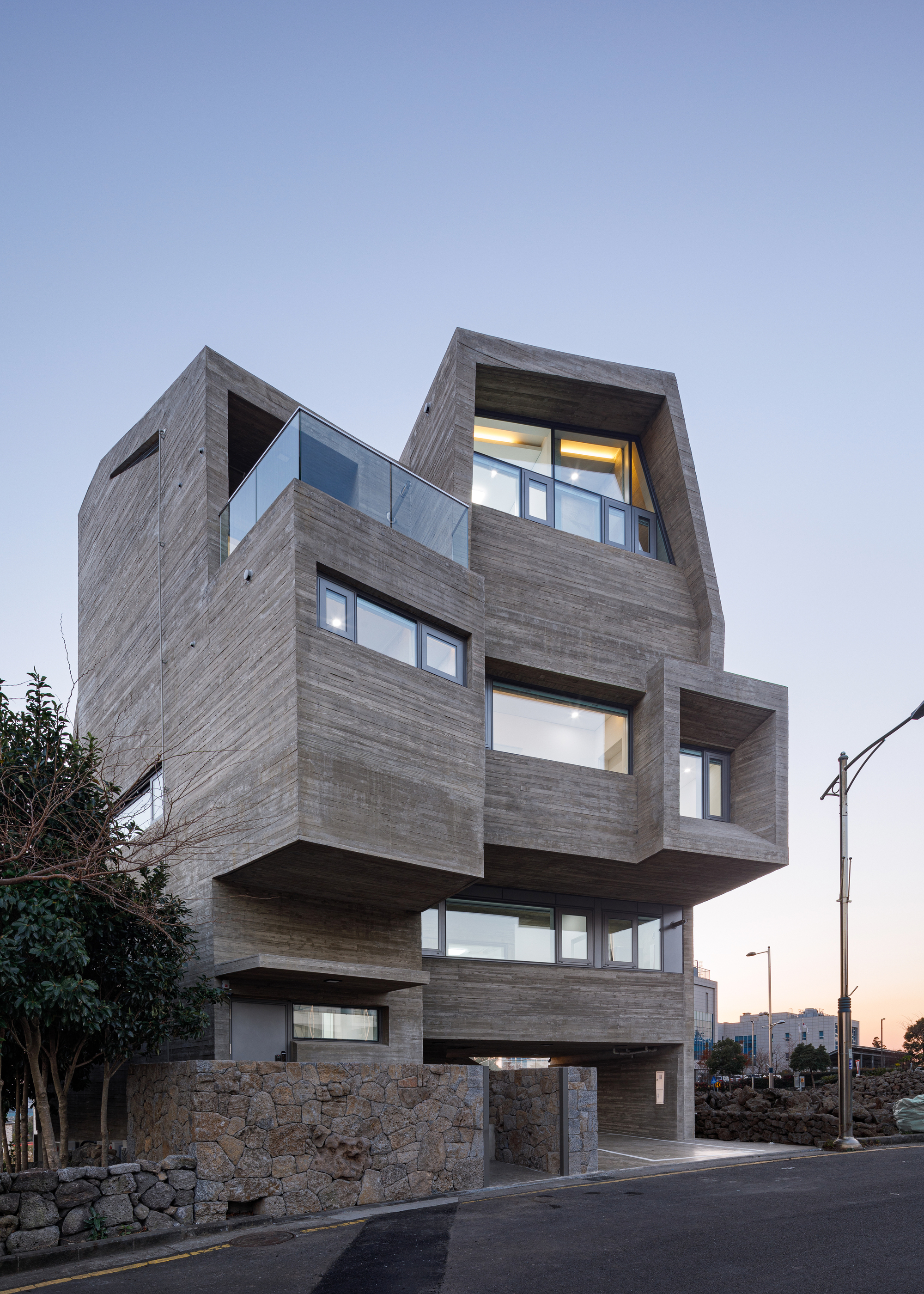 Stacked timber-clad volumes with cantilevered floors and illuminated interiors at twilight beside a residential street