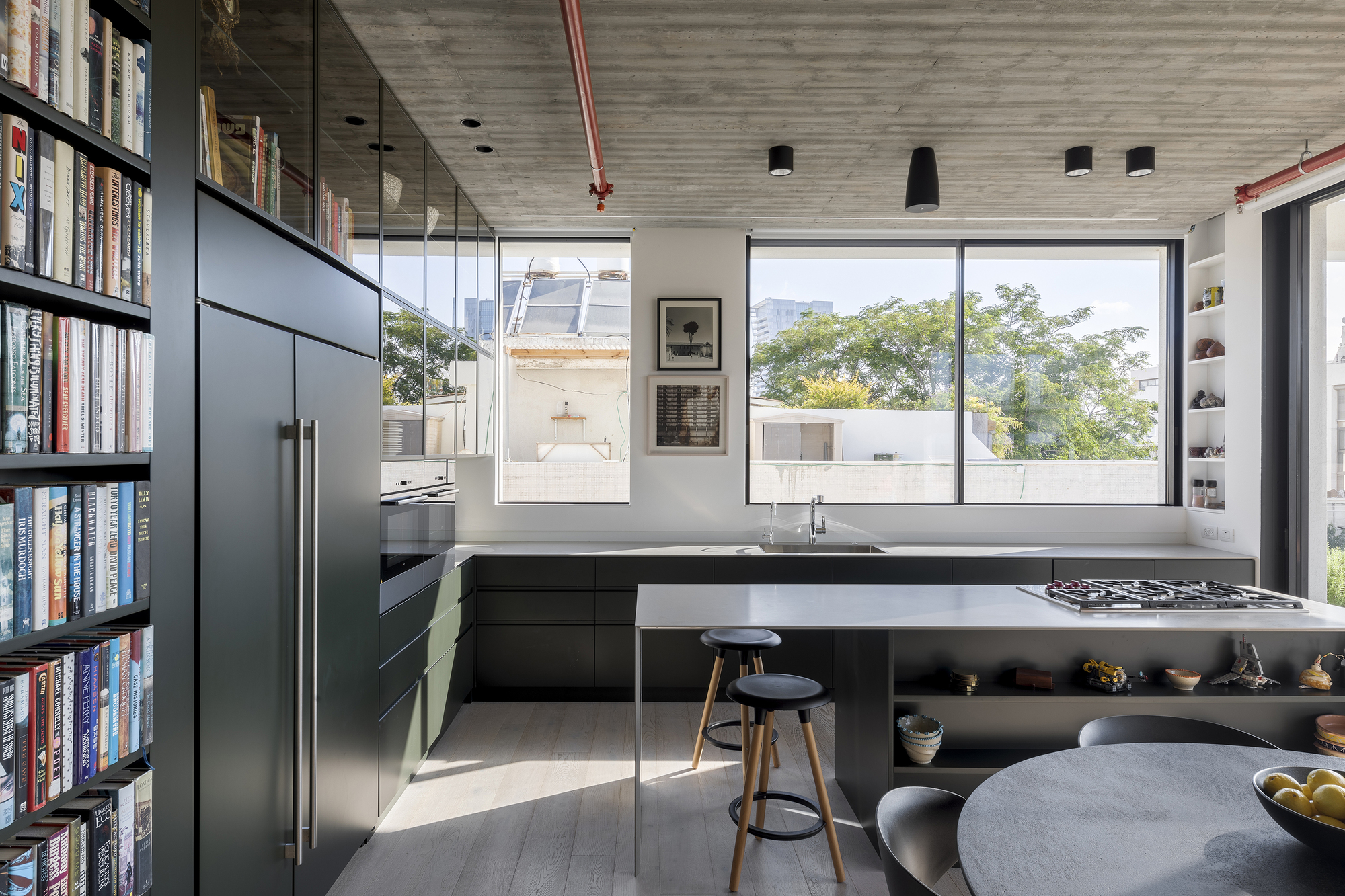 Kitchen island with integrated cooktop beside full-height glazed partition reflecting trees outside in afternoon light