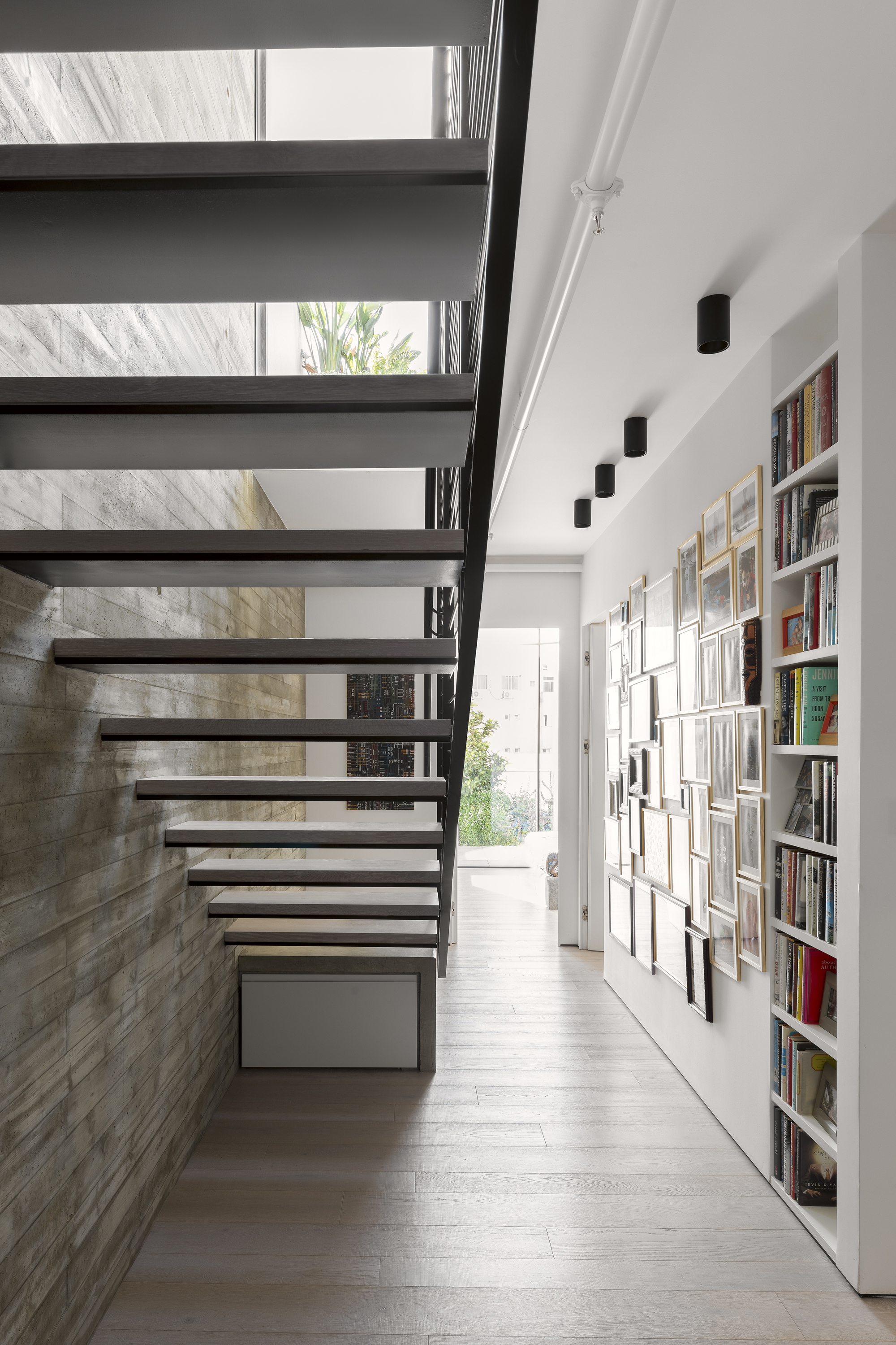 Open tread steel staircase beside board-formed concrete wall with built-in bookshelves and framed artworks