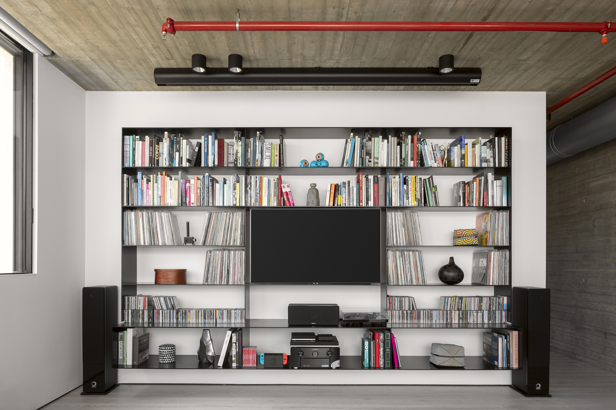 Built-in black shelving unit with television and books below exposed concrete ceiling with red conduit