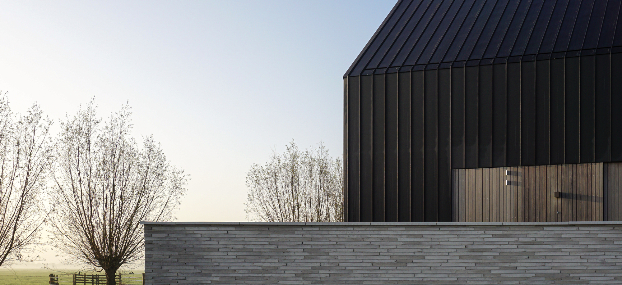 Horizontal stacked stone base wall beneath dark vertical cladding with a tree in misty light