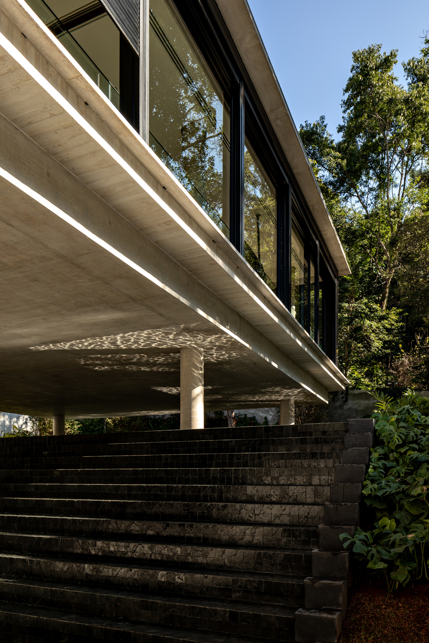 Dark stone staircase ascending beneath a cantilevered concrete soffit with recessed linear lighting at midday