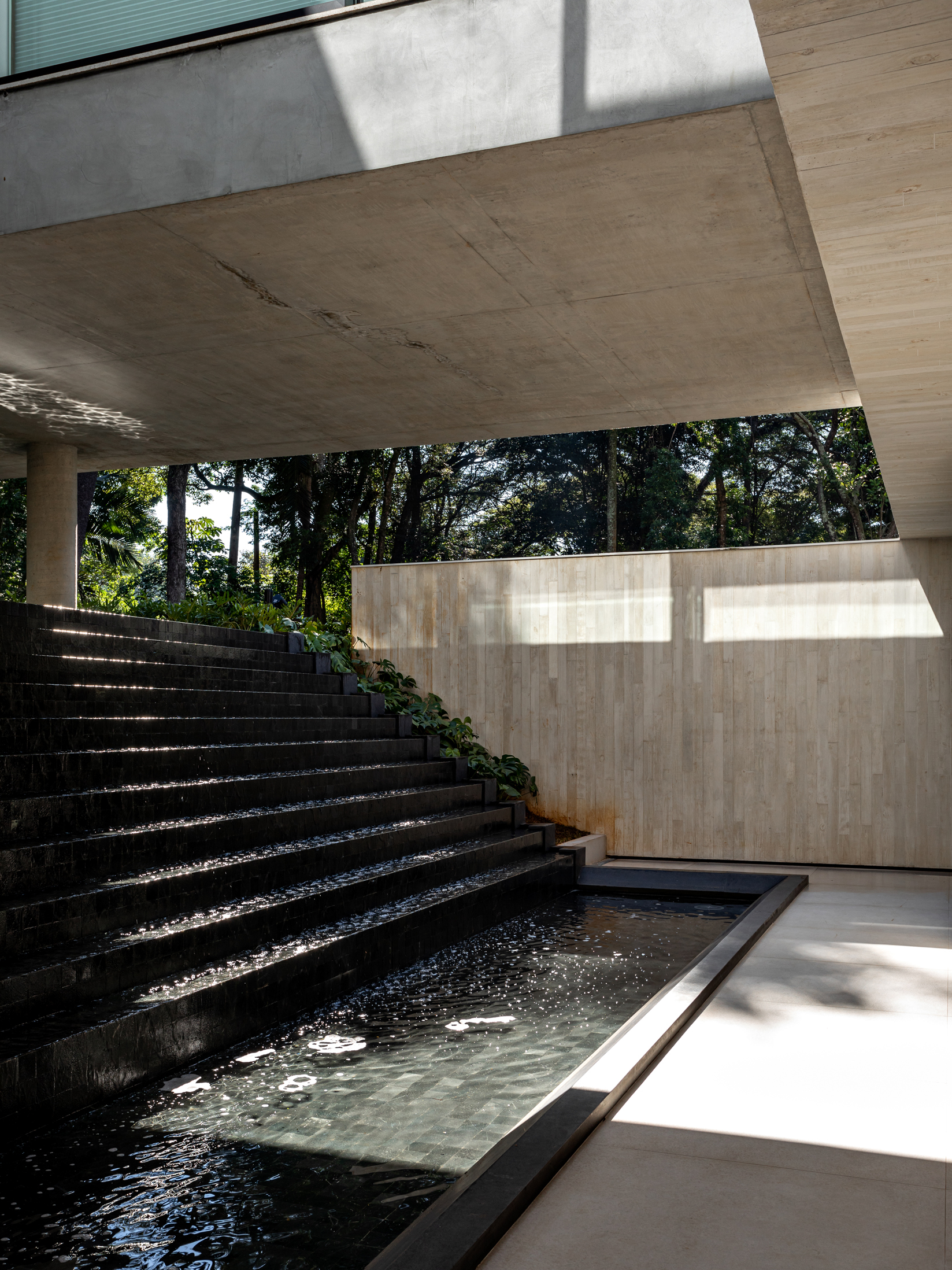 Reflecting pool bordered by stone steps and board-formed concrete walls beneath a deep overhang with filtered light
