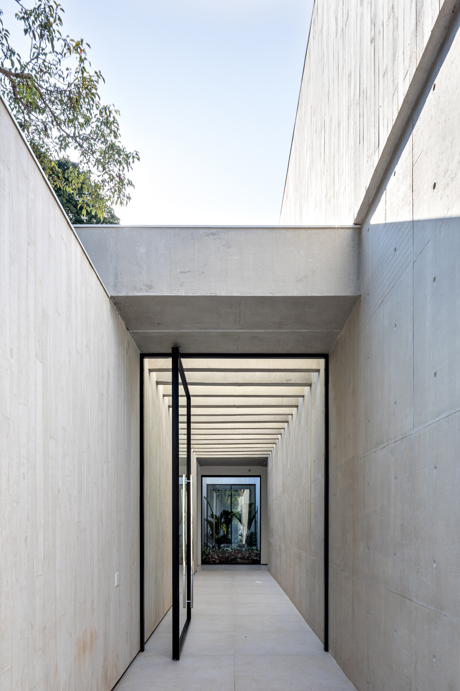 Narrow concrete passageway with open skylight and rhythmic timber beam shadows leading to a glazed door