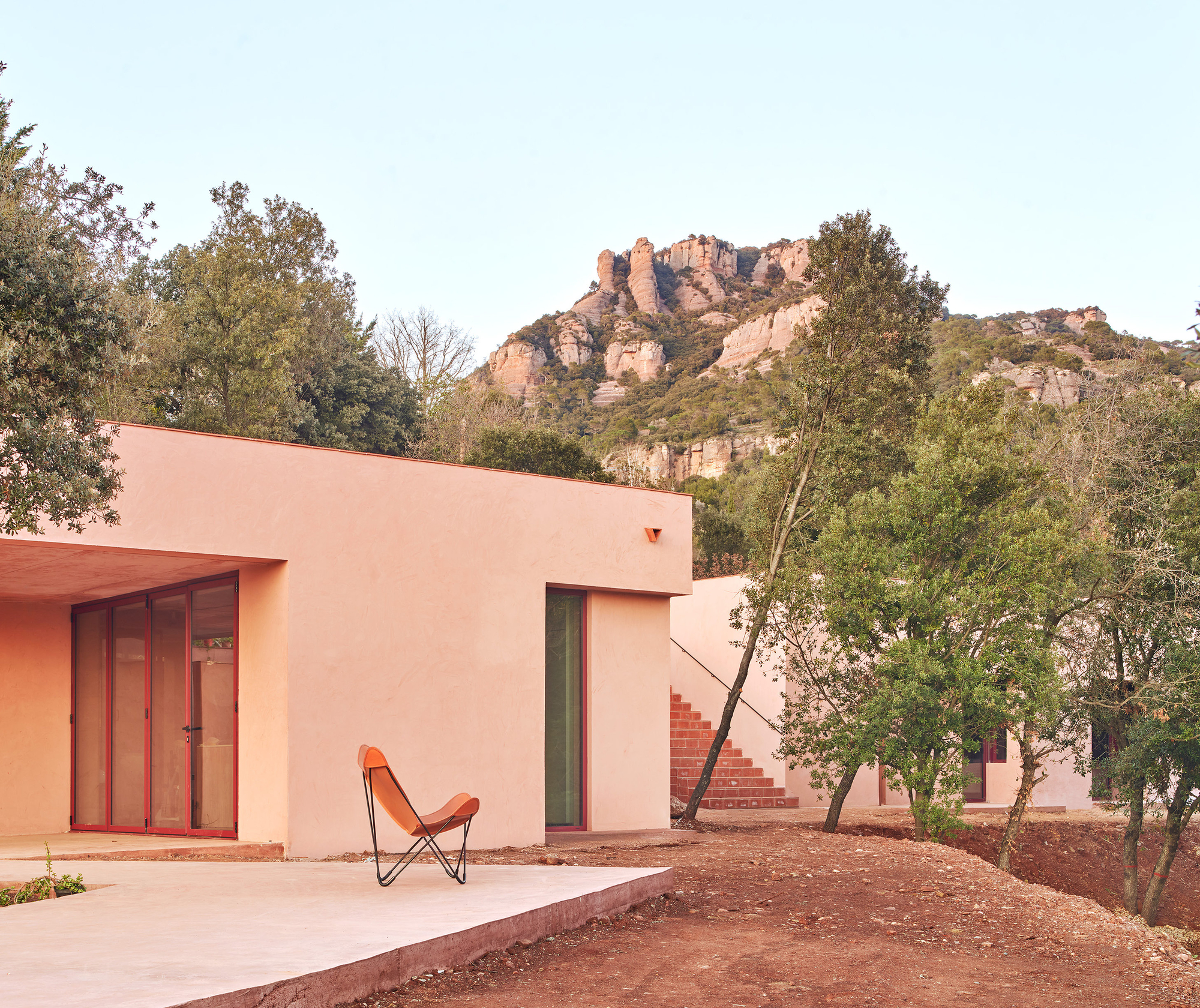 Pink stucco facade with timber-framed glazing and an orange butterfly chair beneath rocky hillside vegetation