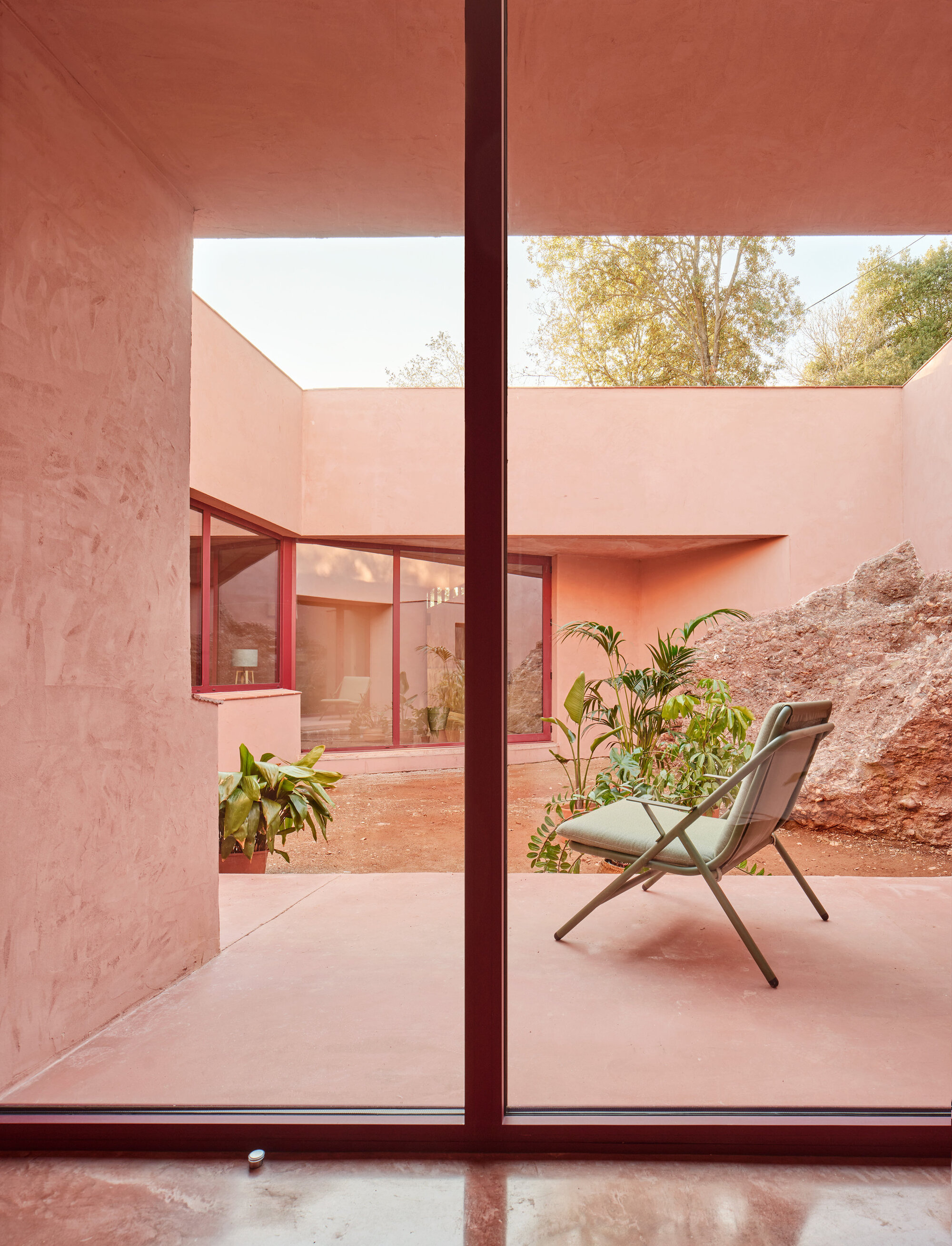 Interior view through floor-to-ceiling glazing onto a pink courtyard with chair and potted palms