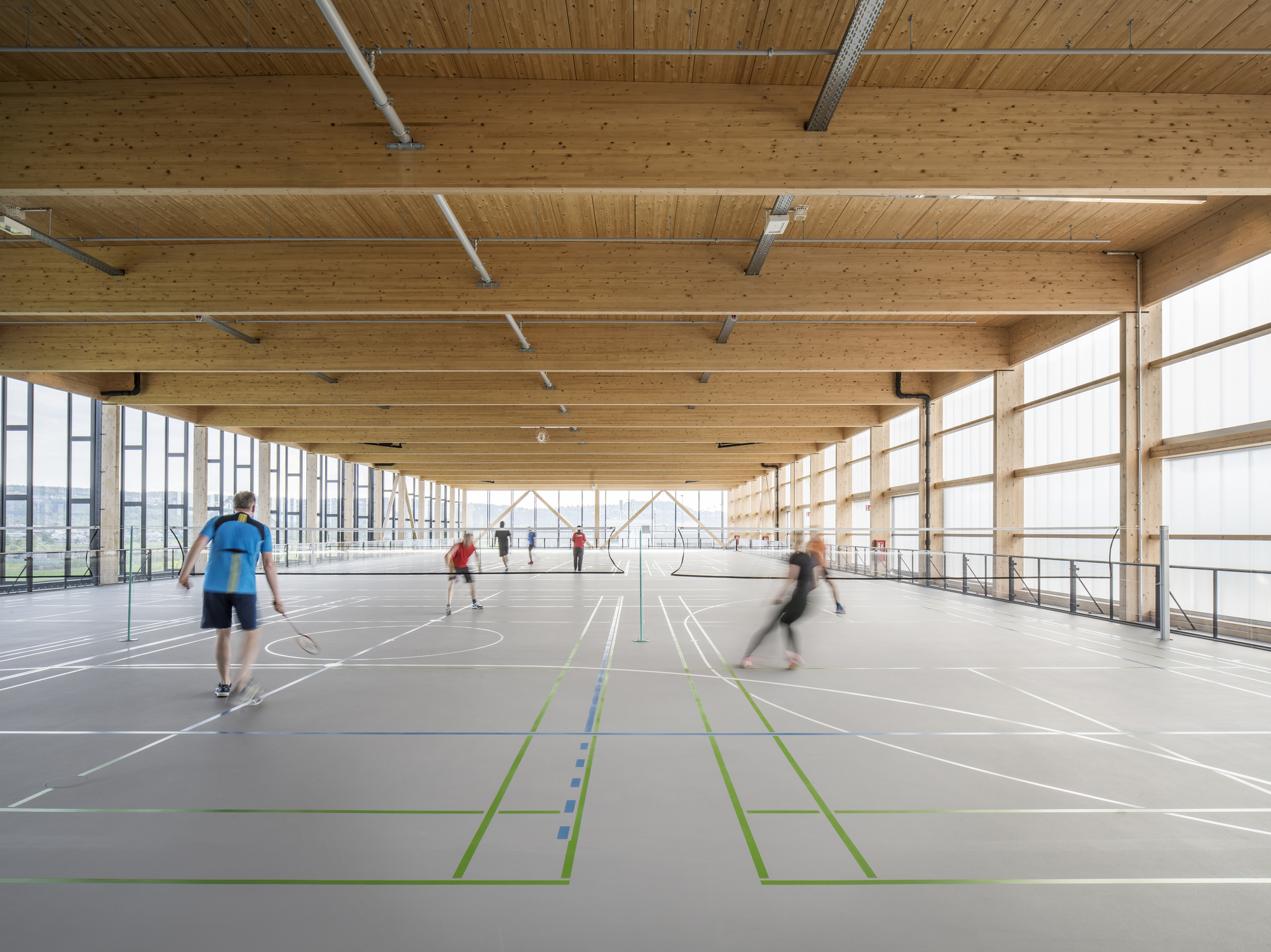Interior sports hall with exposed timber beams and glazed sidewalls with people playing badminton