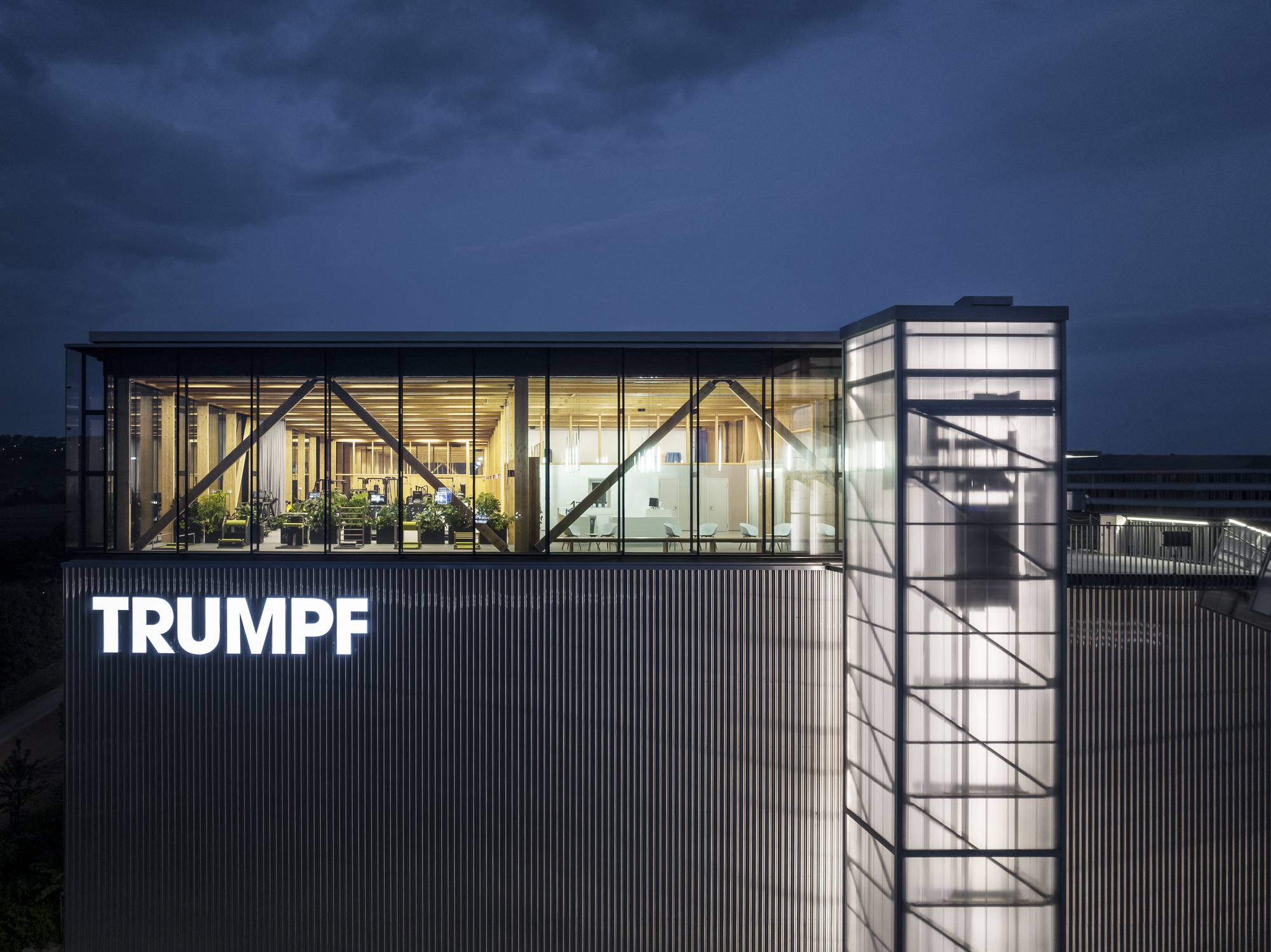 Rooftop glazed pavilion with exposed diagonal bracing and cylindrical stair tower at twilight
