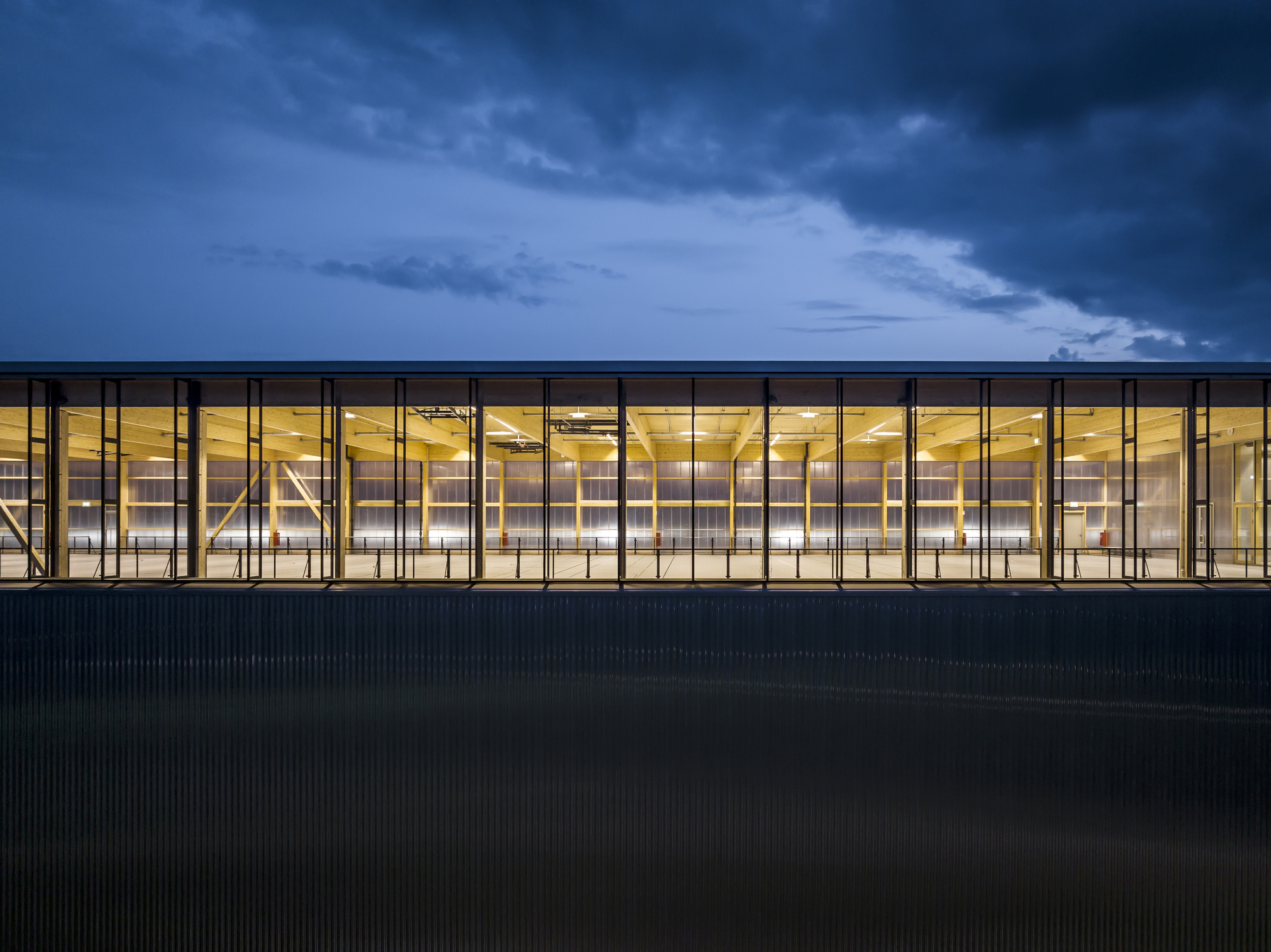 Continuous glass facade with timber ceiling visible inside under dark evening sky