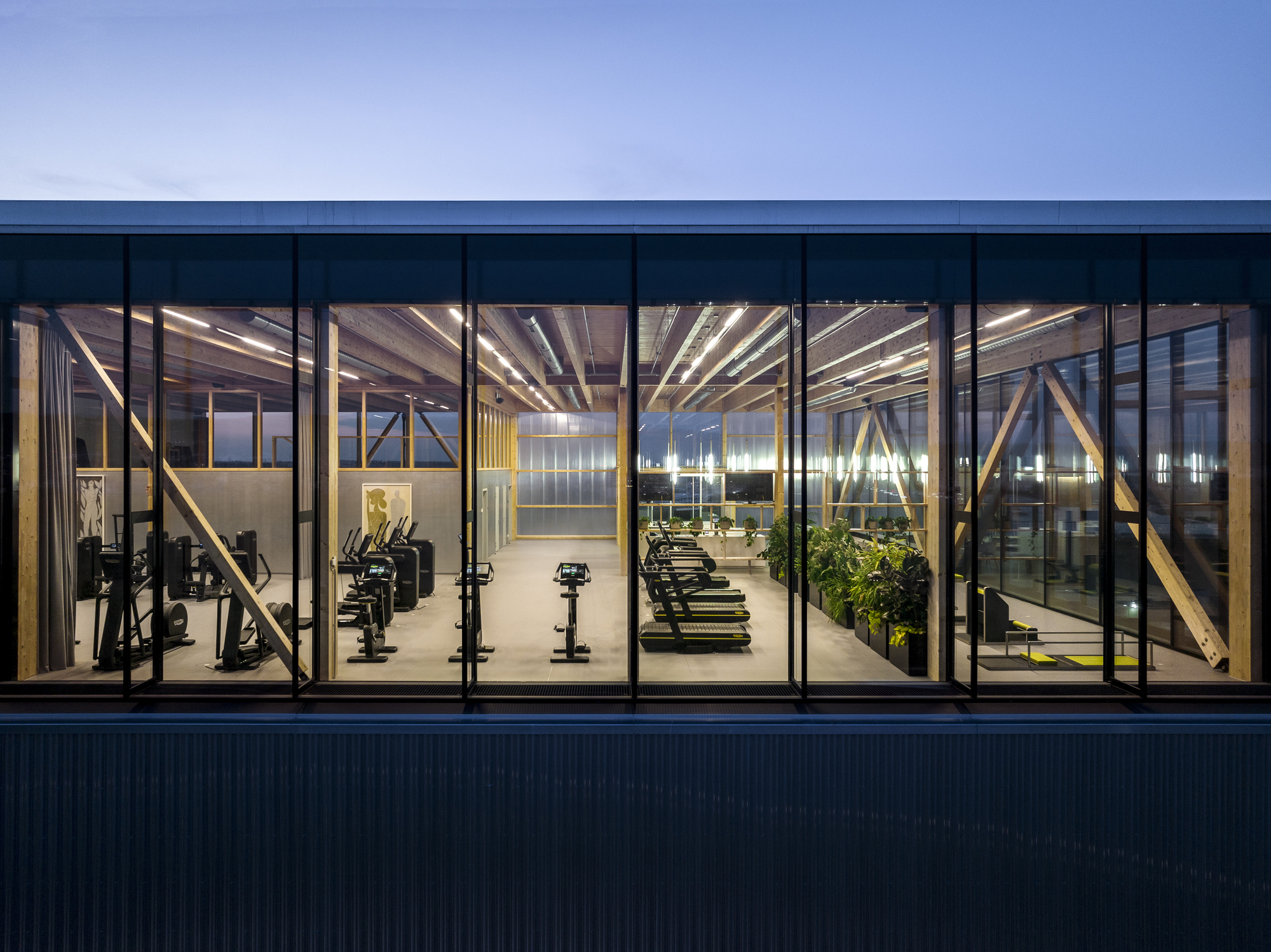 Exterior view through floor-to-ceiling windows revealing gym equipment and planted pots at dusk