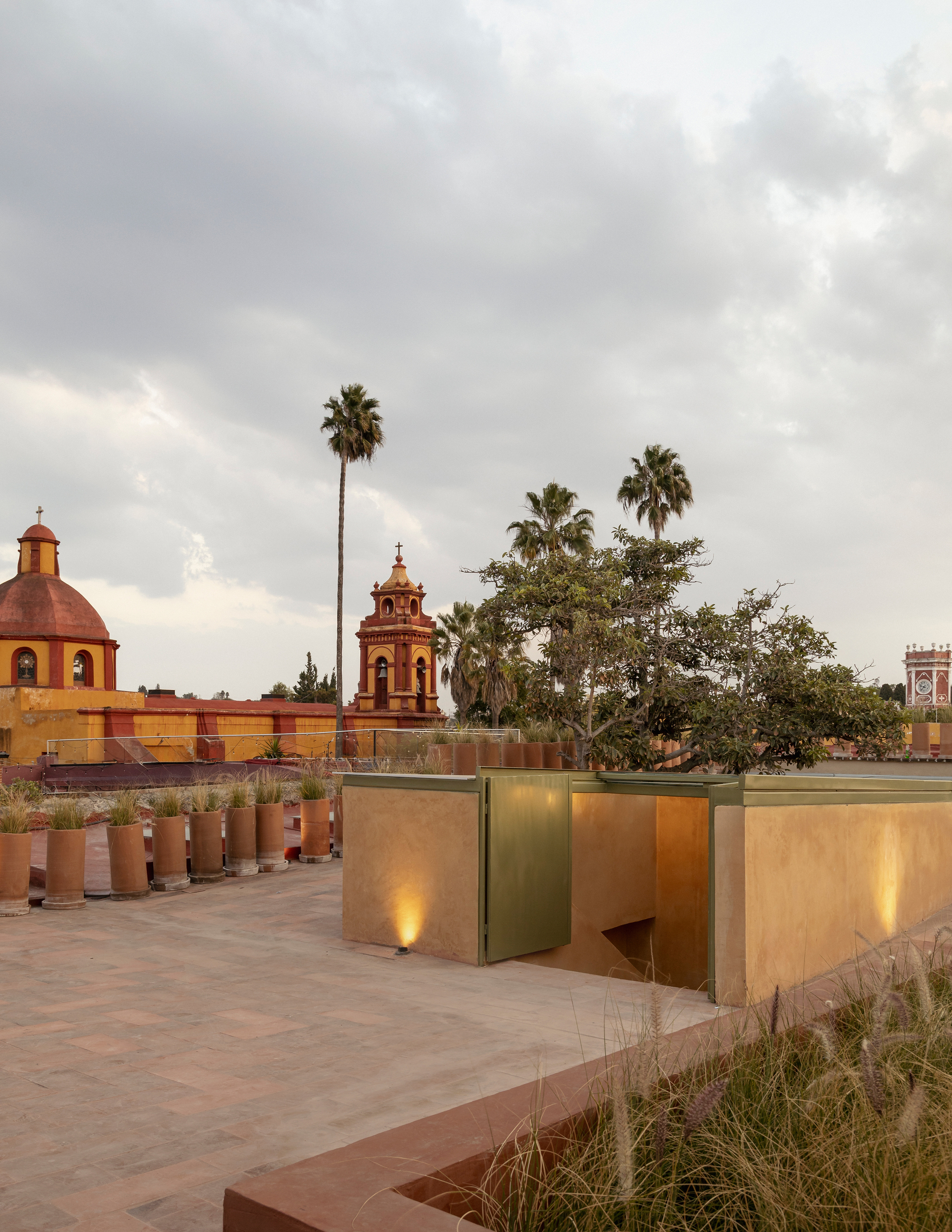 Rooftop terrace with brass-clad volumes and planted beds under cloudy skies with palms and church towers beyond
