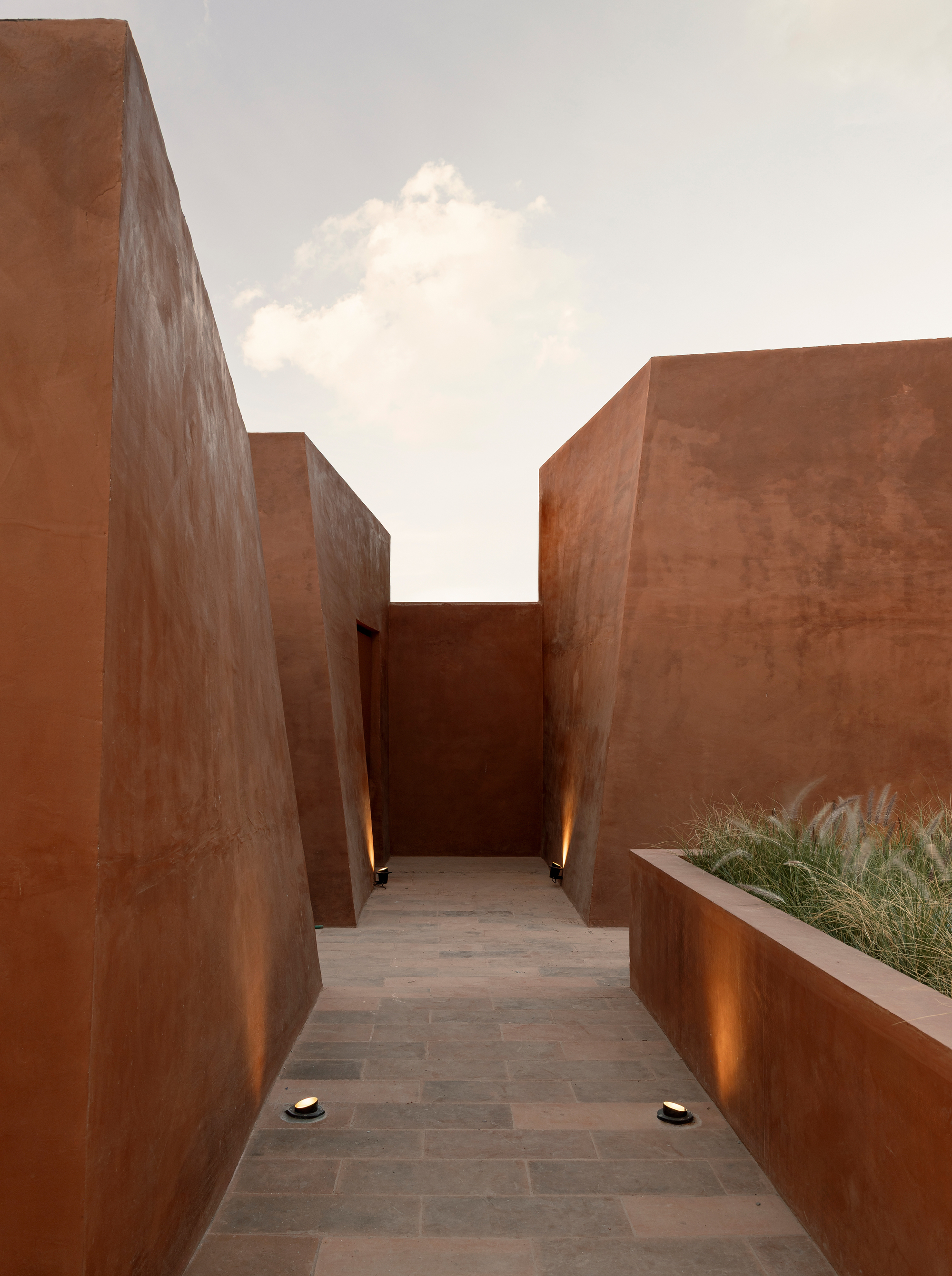 Passageway between corten steel walls with planted bed and uplighting beneath a white cloud-filled sky