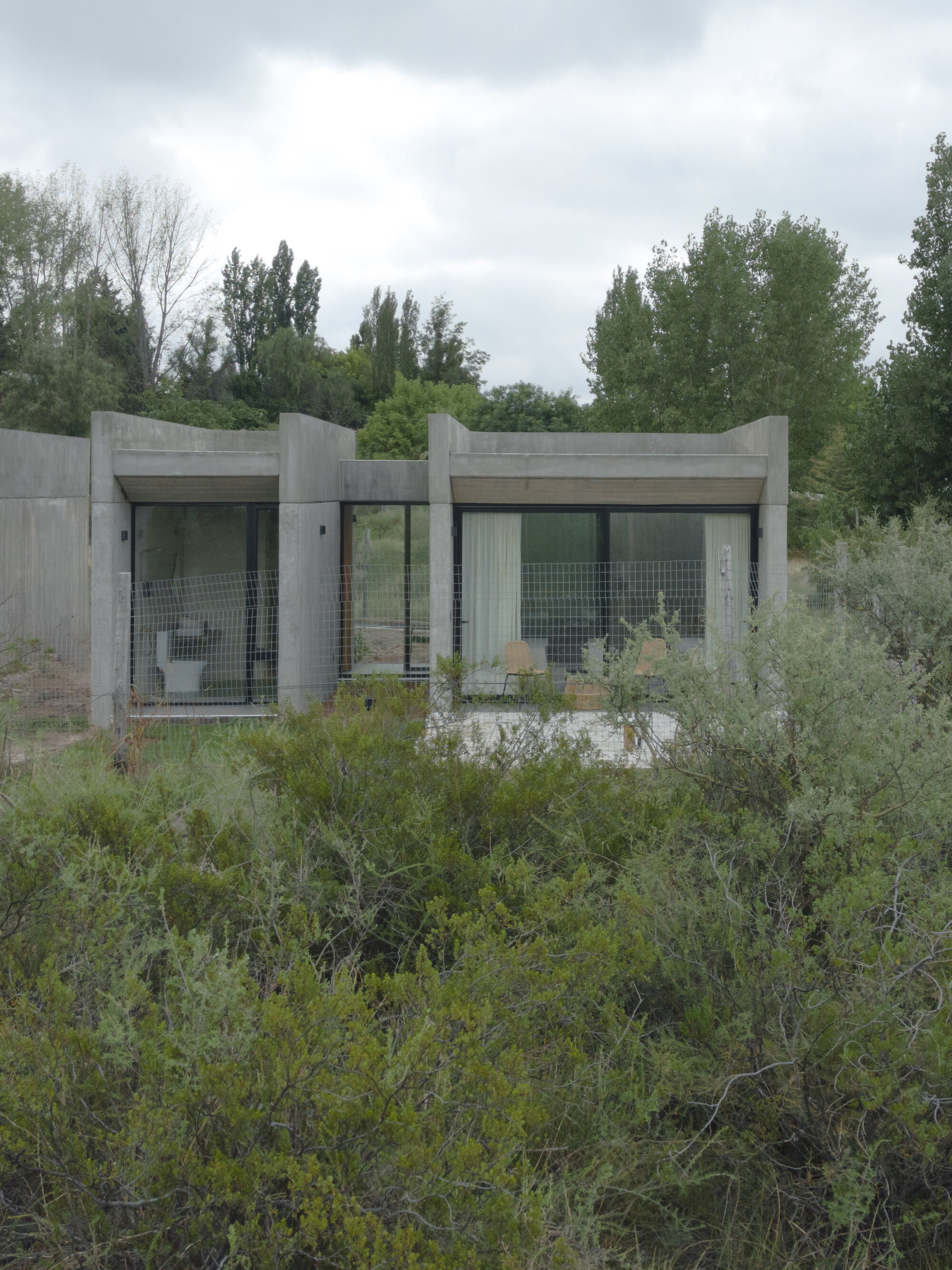 Two-lobed concrete structure with glazed openings surrounded by dense scrubland under cloudy skies