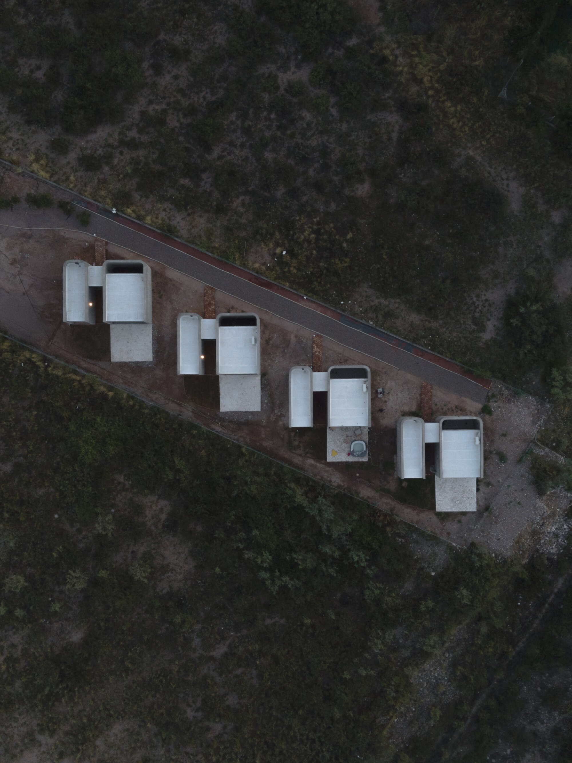 Aerial view of four white rectangular housing units aligned along a roadway in scrubland at dusk
