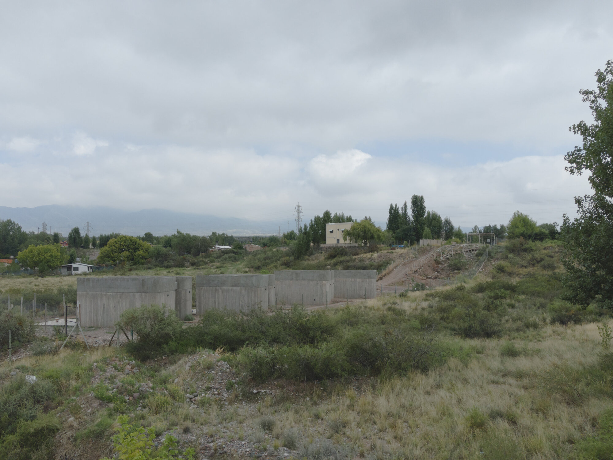 Board-formed concrete retaining wall set into a hillside with scrubland vegetation under overcast sky