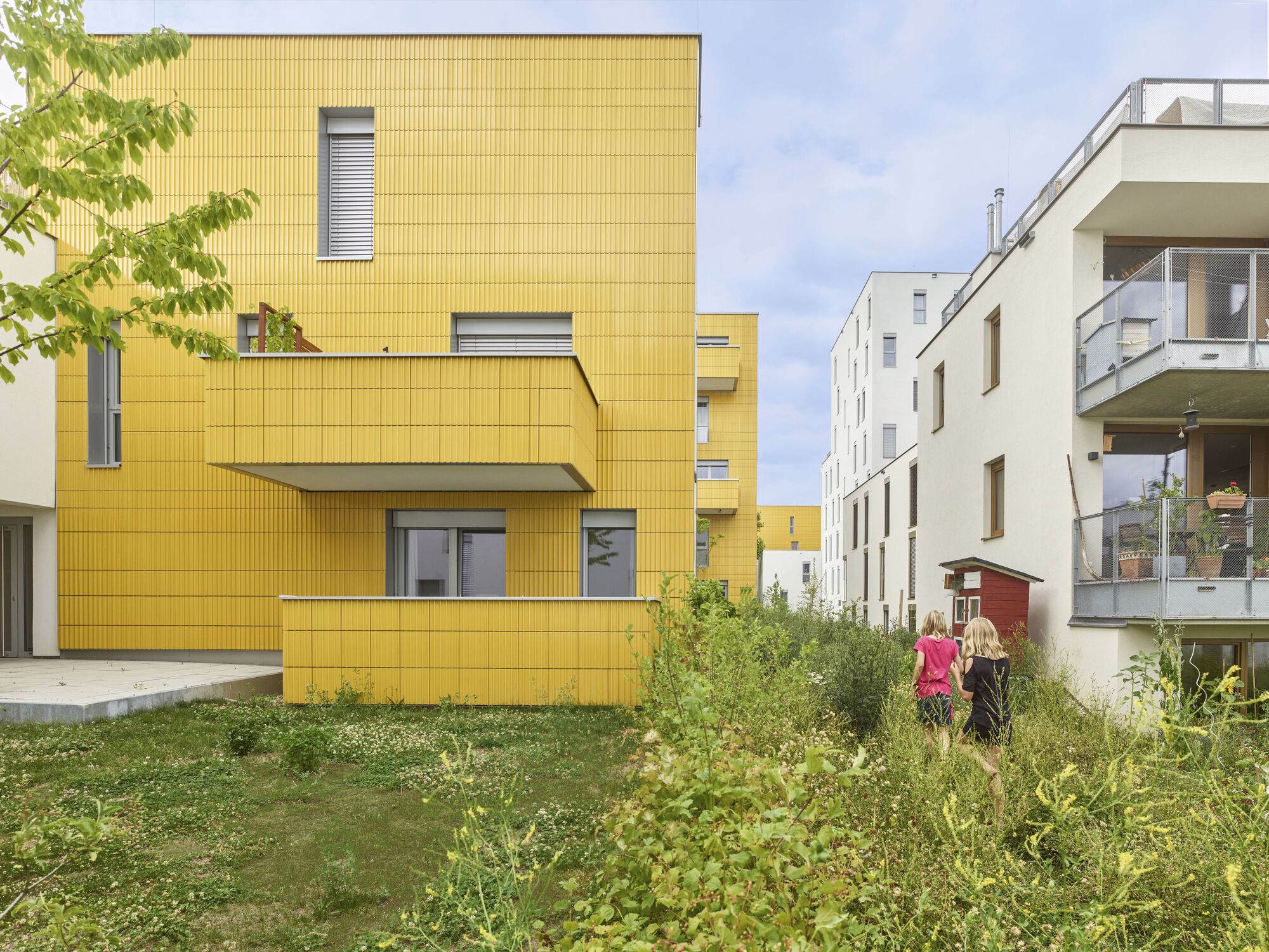 Yellow corrugated metal facade with cantilevered balcony and green planting in the courtyard below