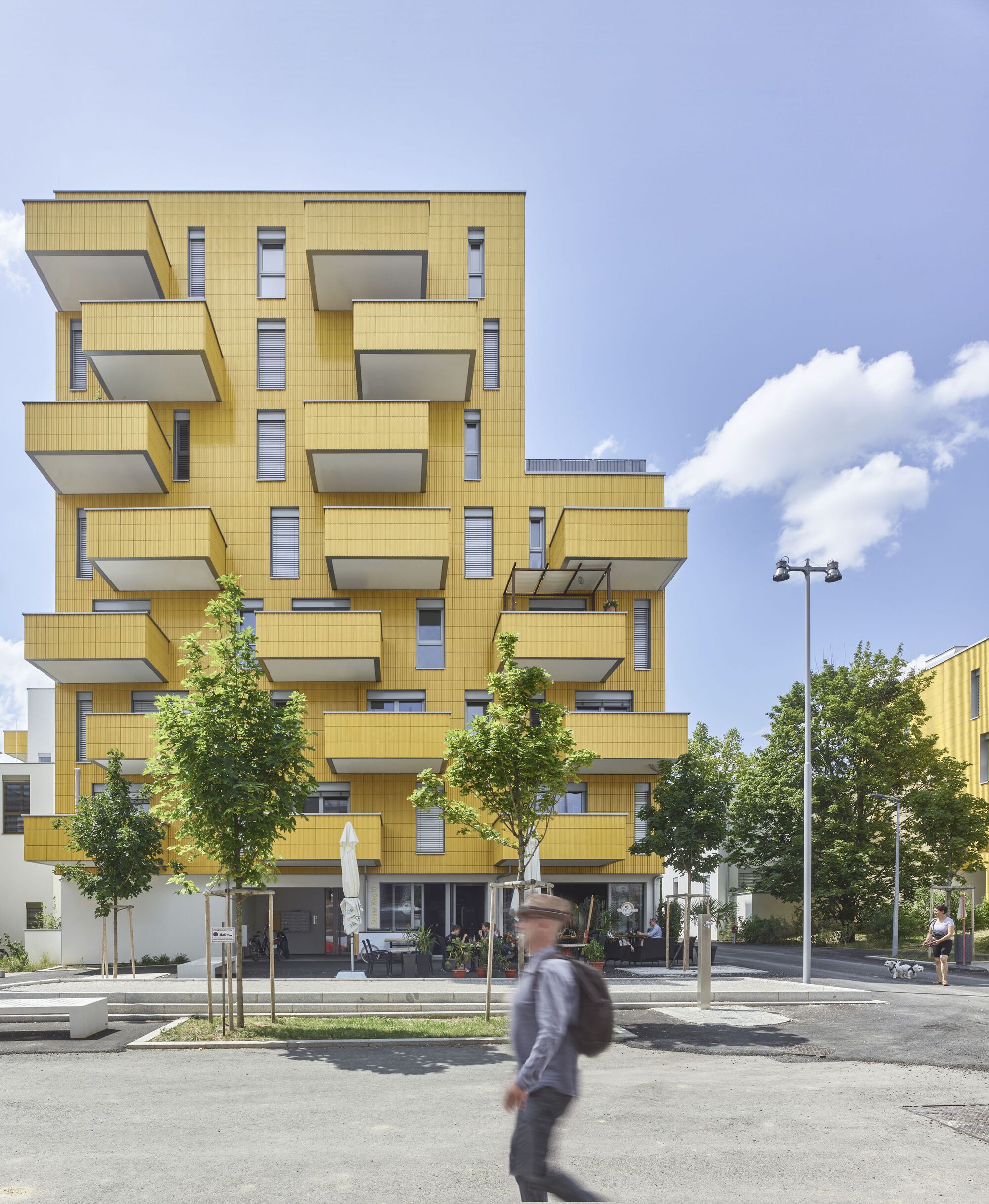 Yellow corrugated metal facade with staggered balconies framed by street trees and a passing pedestrian
