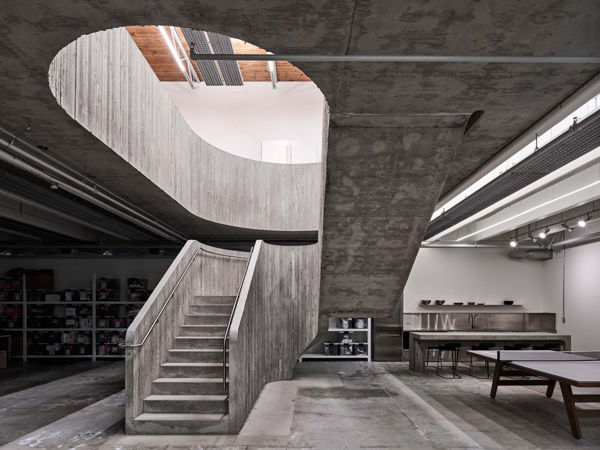 Curved concrete staircase with board-formed walls rising through an oval ceiling opening to natural light