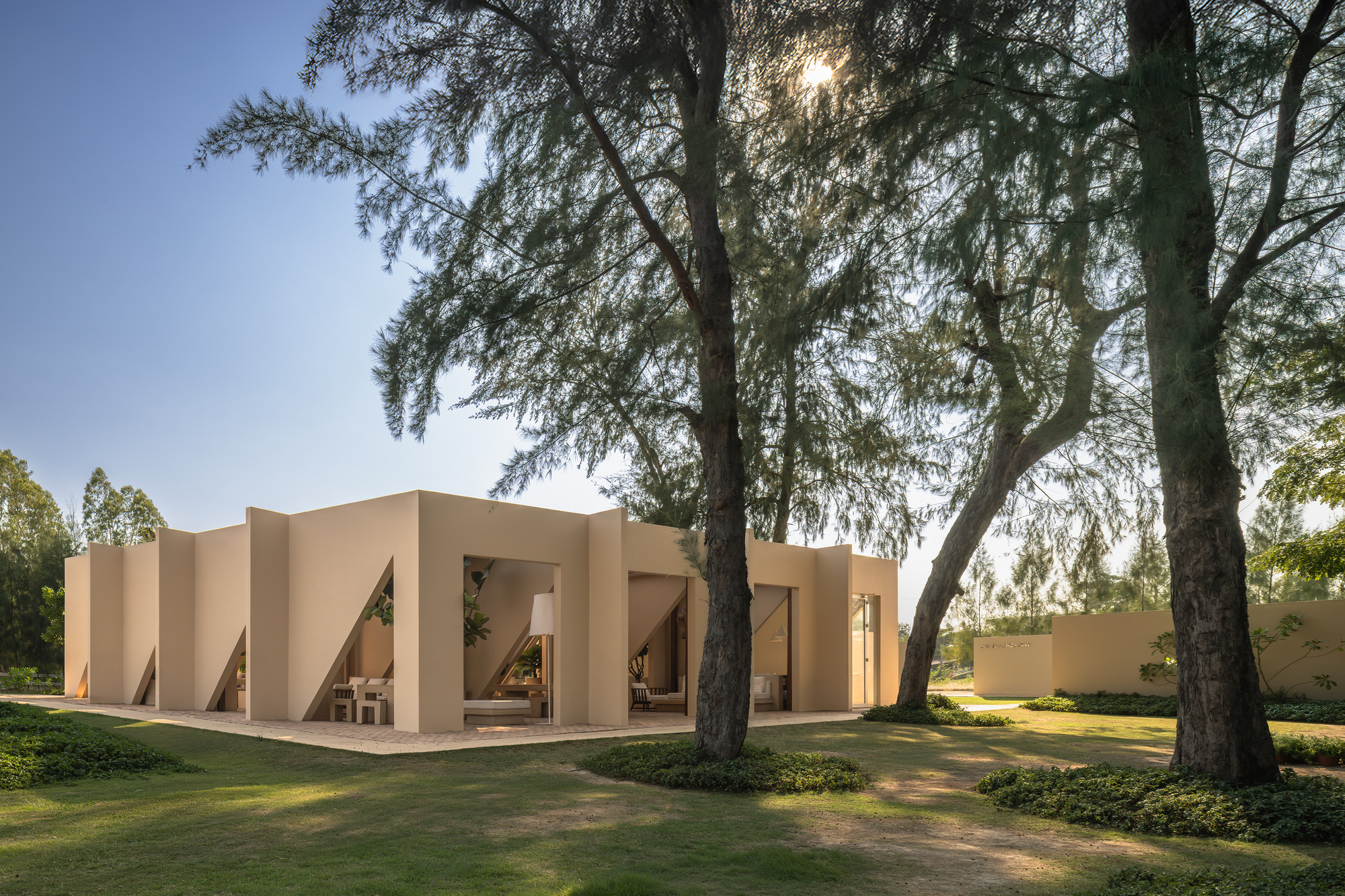 Folded facade with angled openings framing a covered porch beneath tall trees in afternoon light