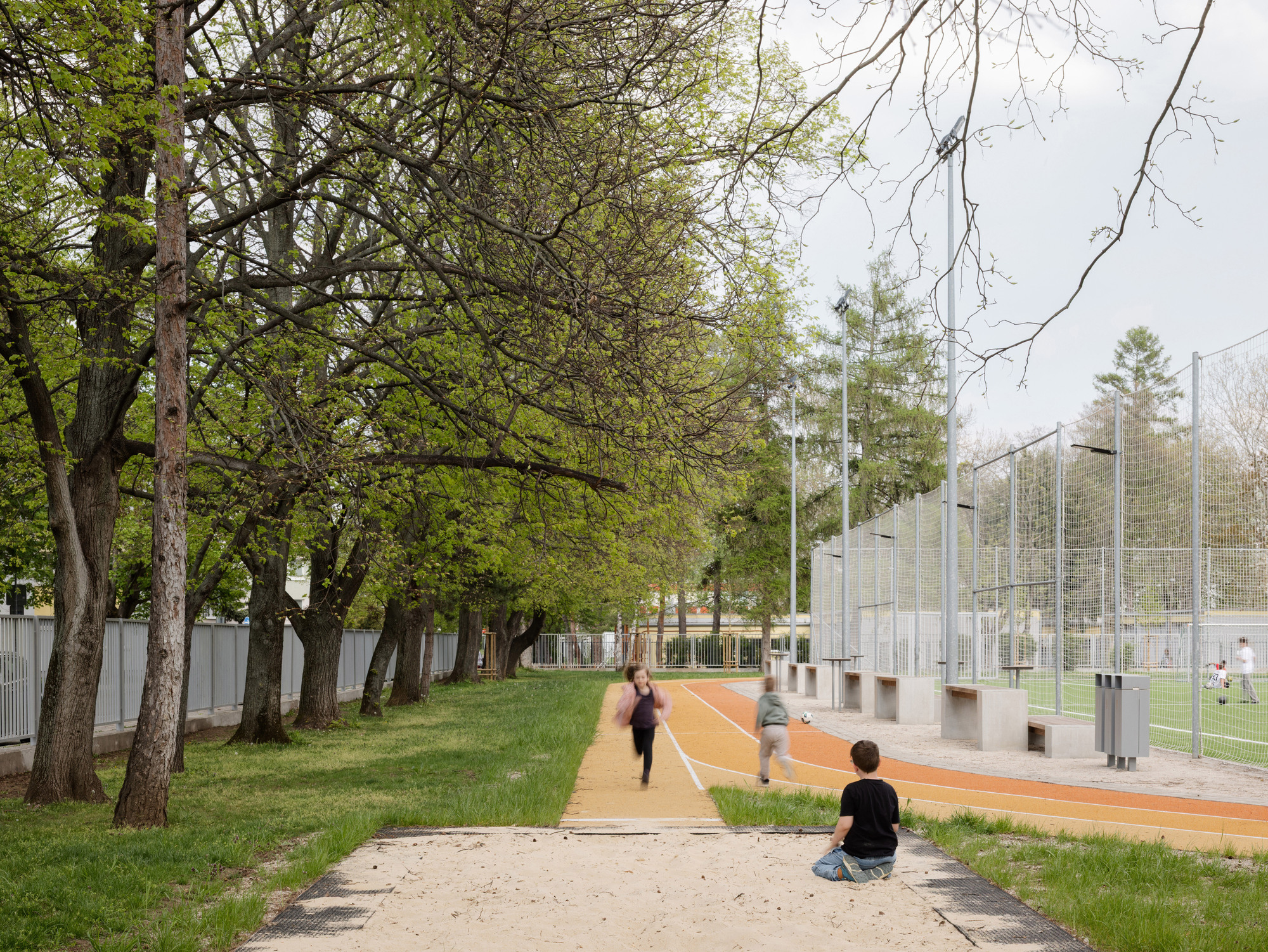 Tree-lined path beside the sports court with figures running in dappled shade