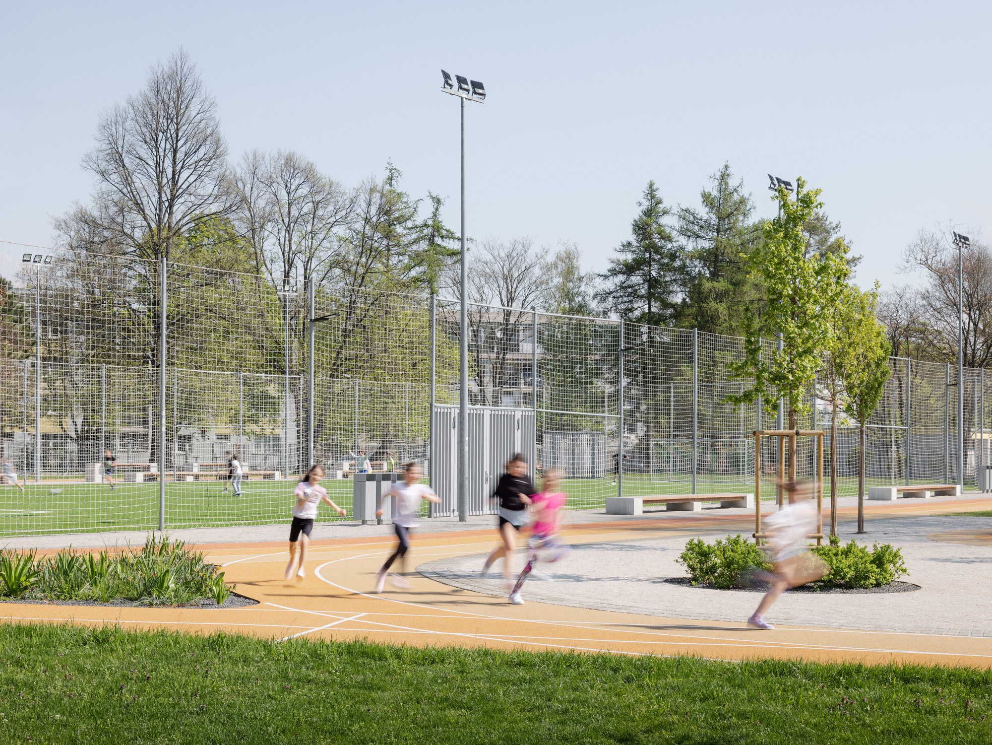 Children running across a curved plaza with planted beds and mesh sports enclosure beyond