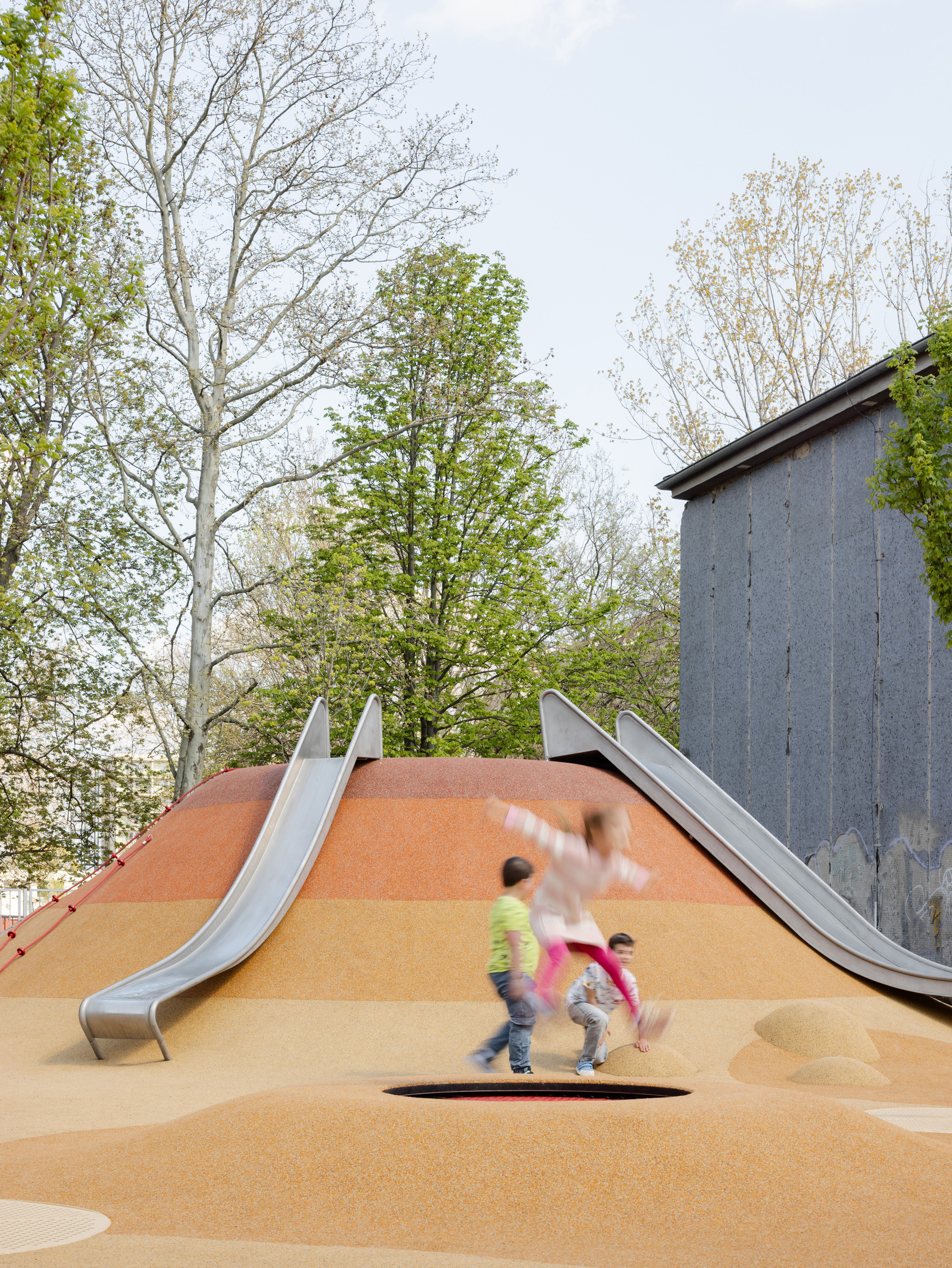 Contoured play mound with stainless steel slides and children running near concrete structure under cloudy sky
