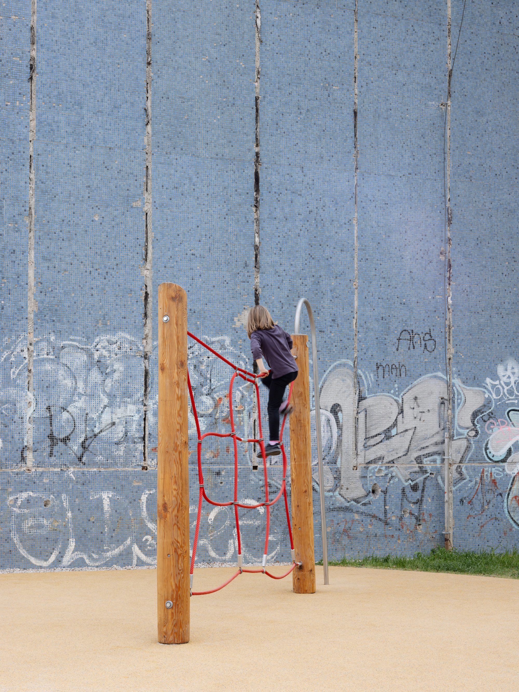 Child climbing red rope net structure on timber posts against a weathered blue tile wall