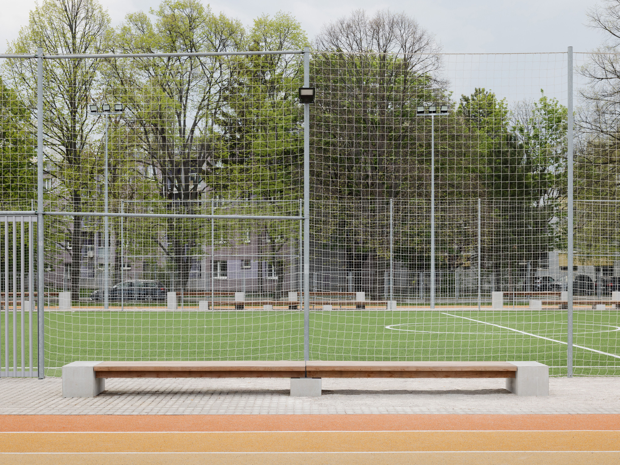 Timber bench with concrete endcaps facing a synthetic turf sports pitch enclosed by wire fencing