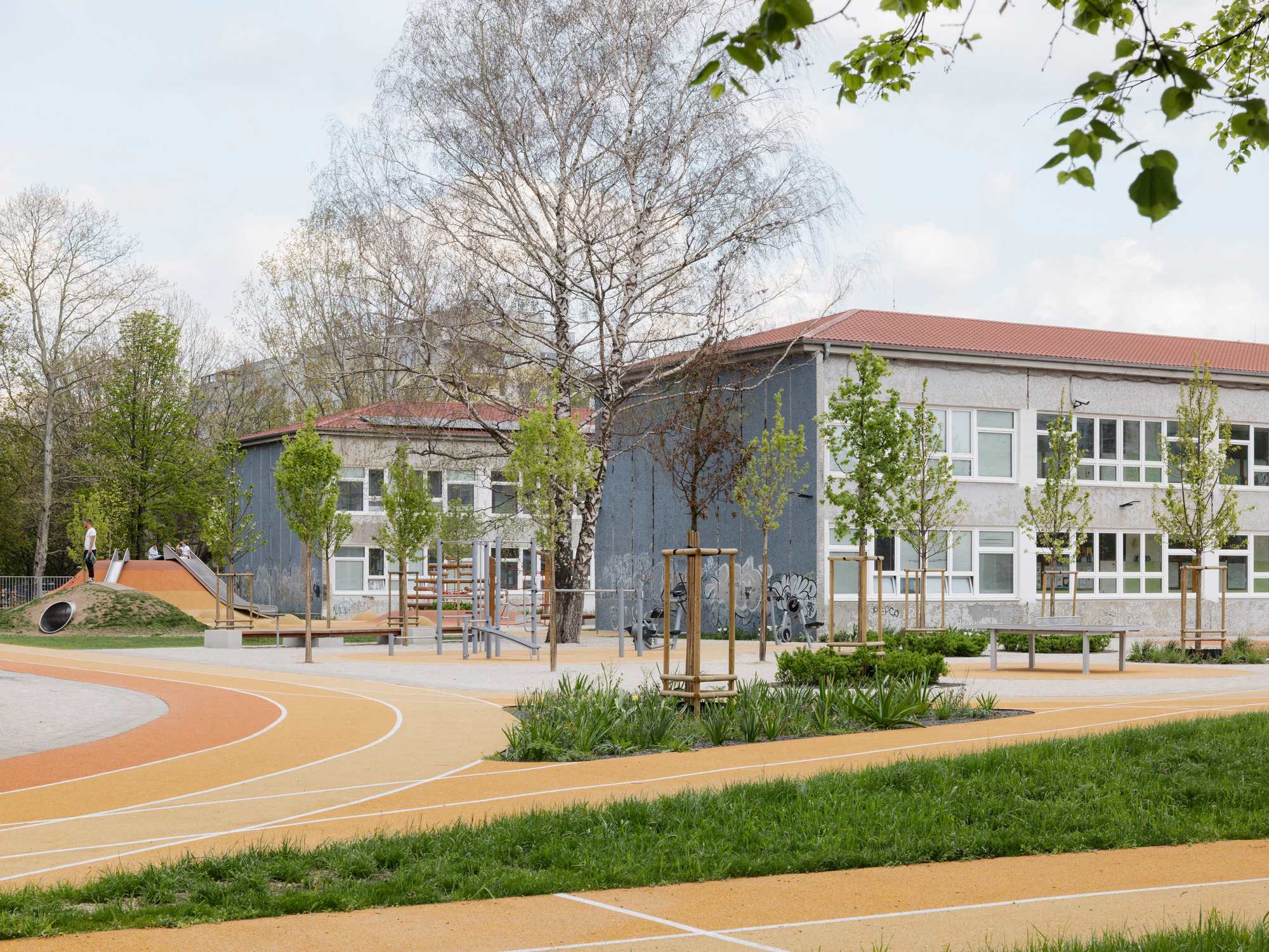 Schoolyard with orange rubber surfacing and planted beds between two gabled buildings under overcast skies