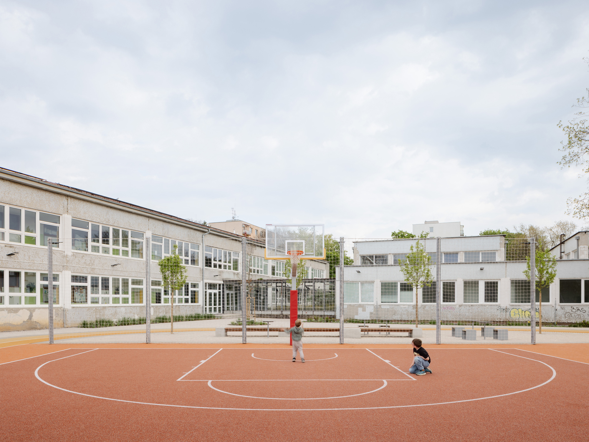 Basketball court with orange rubber surface where two children play beneath a cloudy sky