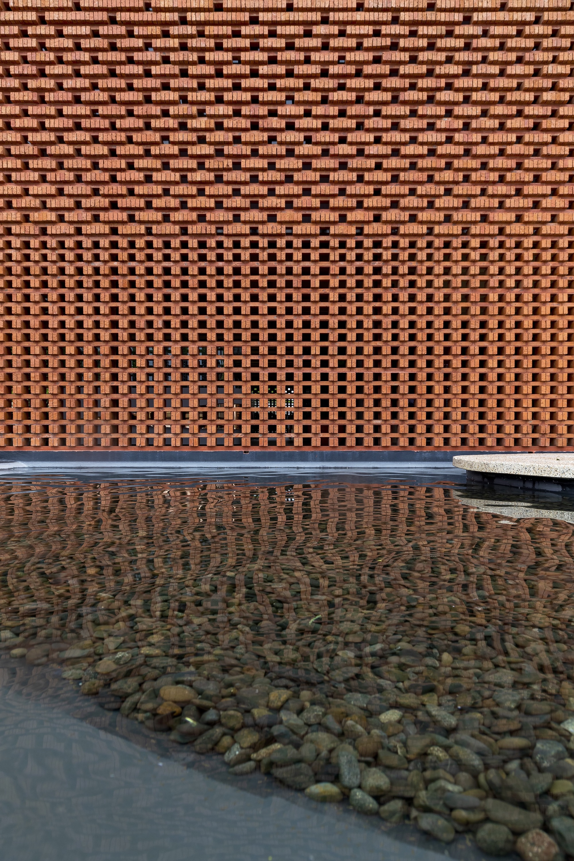 Perforated brick screen wall with varying aperture patterns reflected in shallow water pool with river stones