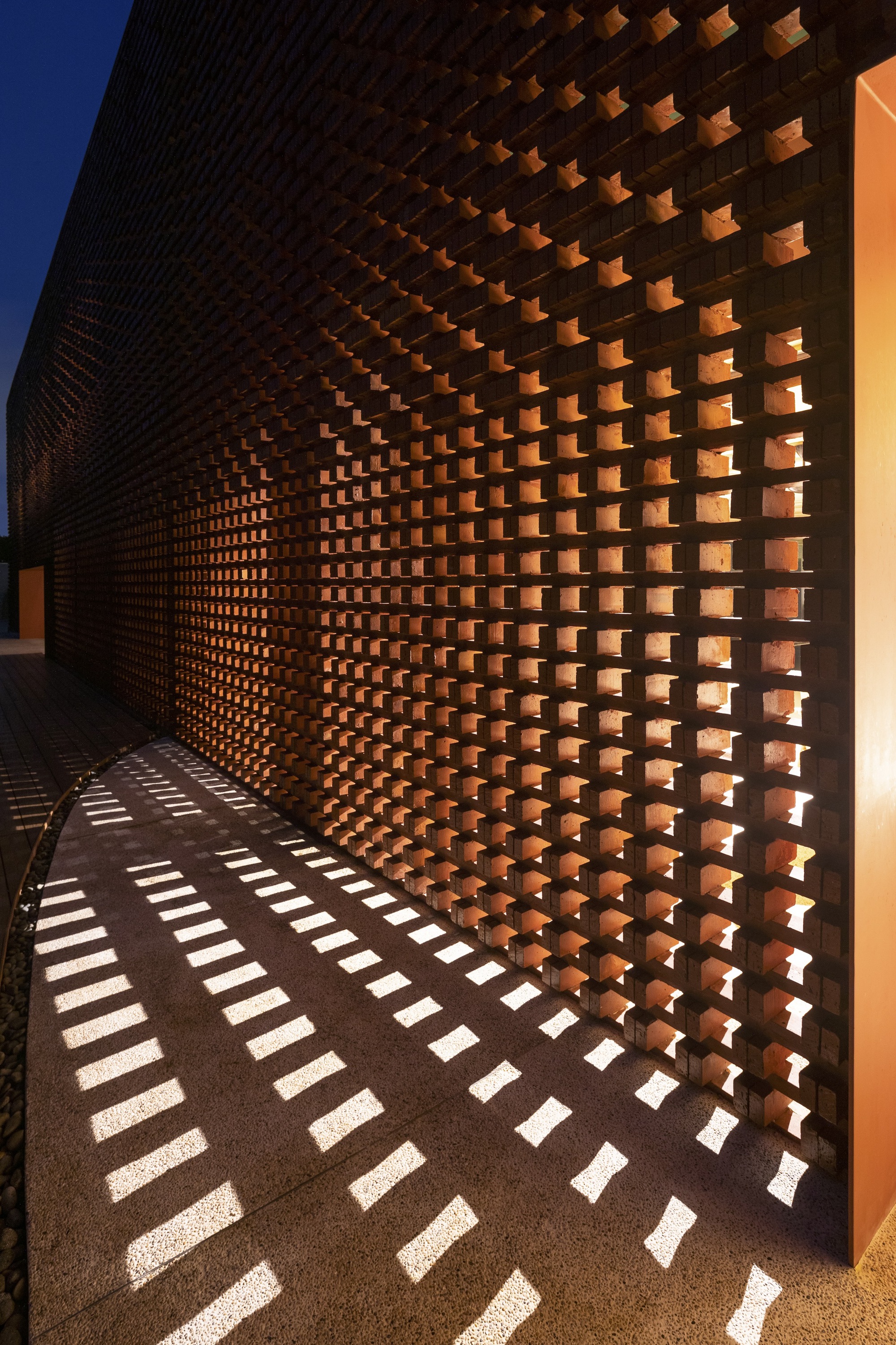 Curved perforated brick wall casting dappled shadows on the ground at dusk