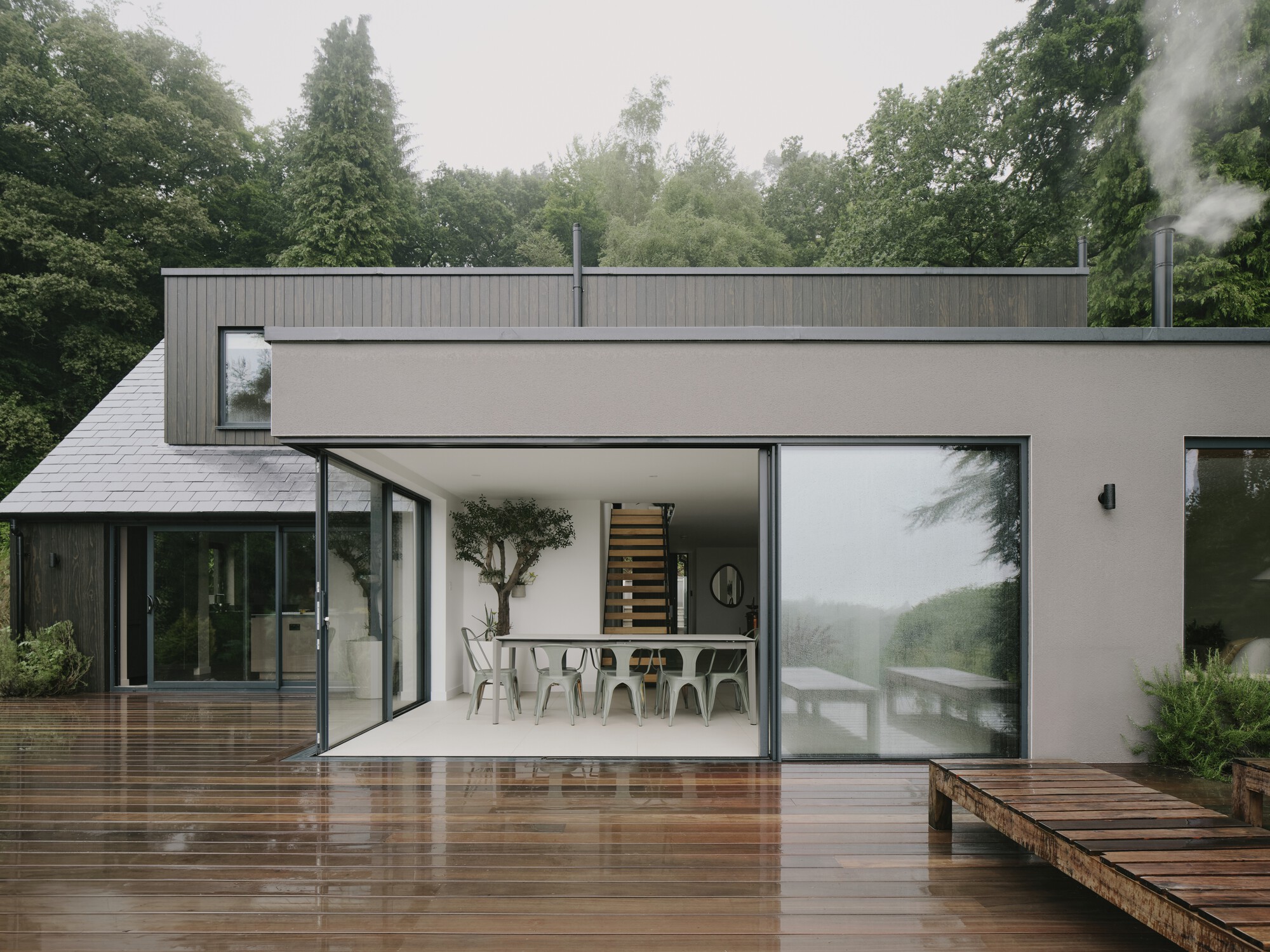Open glass doors connecting the dining area to a wet timber deck under misty skies