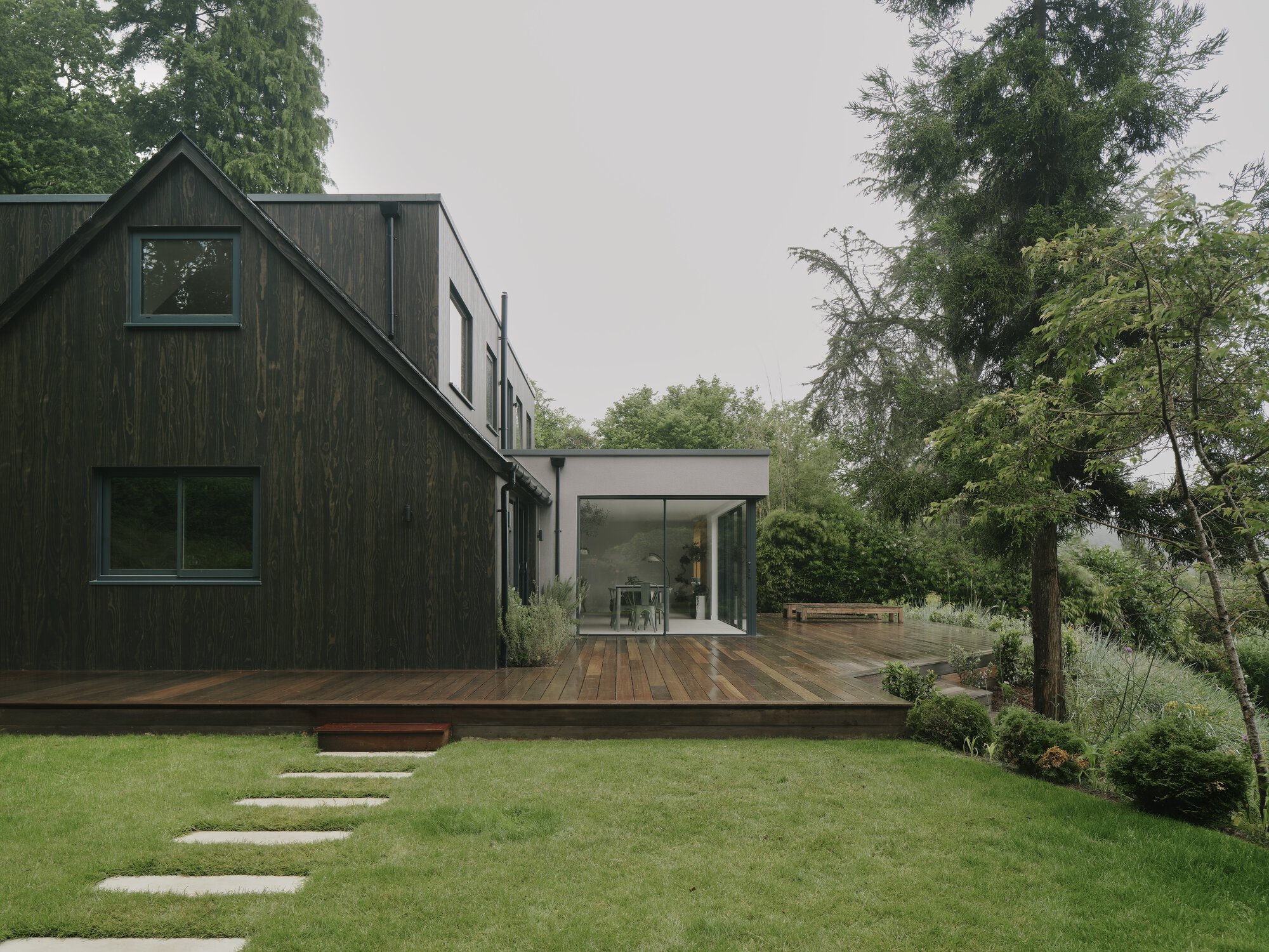 Blackened timber gable volume and glazed extension overlooking a stepped stone path through lawn