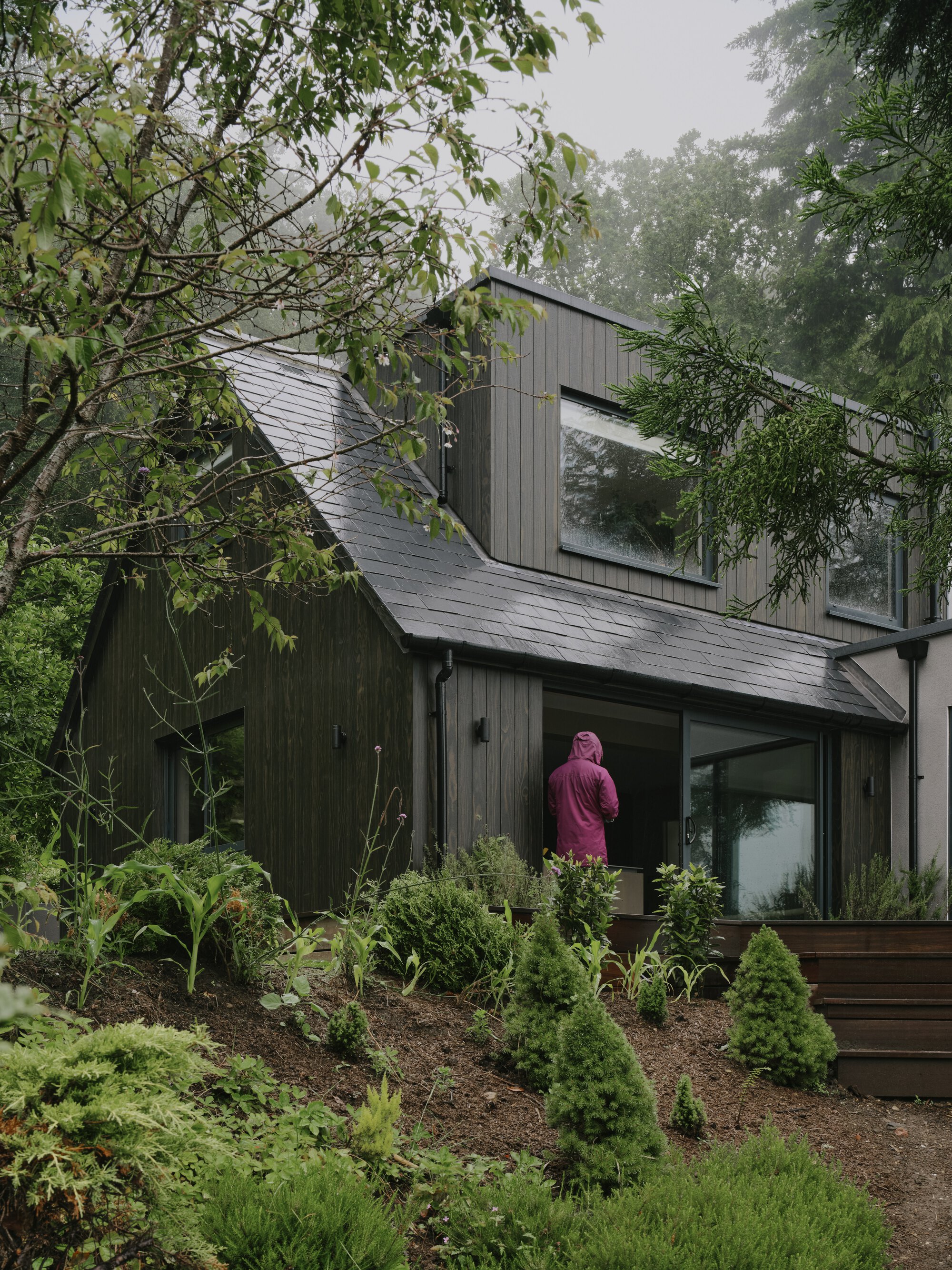 Figure in pink robe on planted slope below the dark timber and slate facade