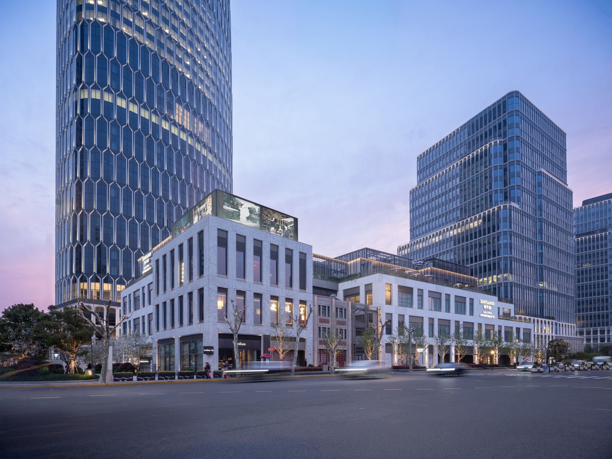 Street view of white framed podium and glass towers at dusk with blooming trees along the sidewalk