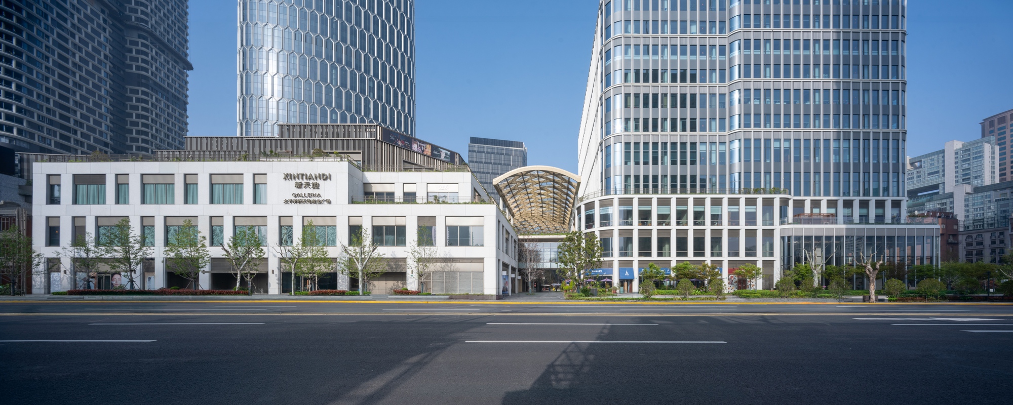Panoramic view across the empty street showing white podium and woven canopy between glass office towers
