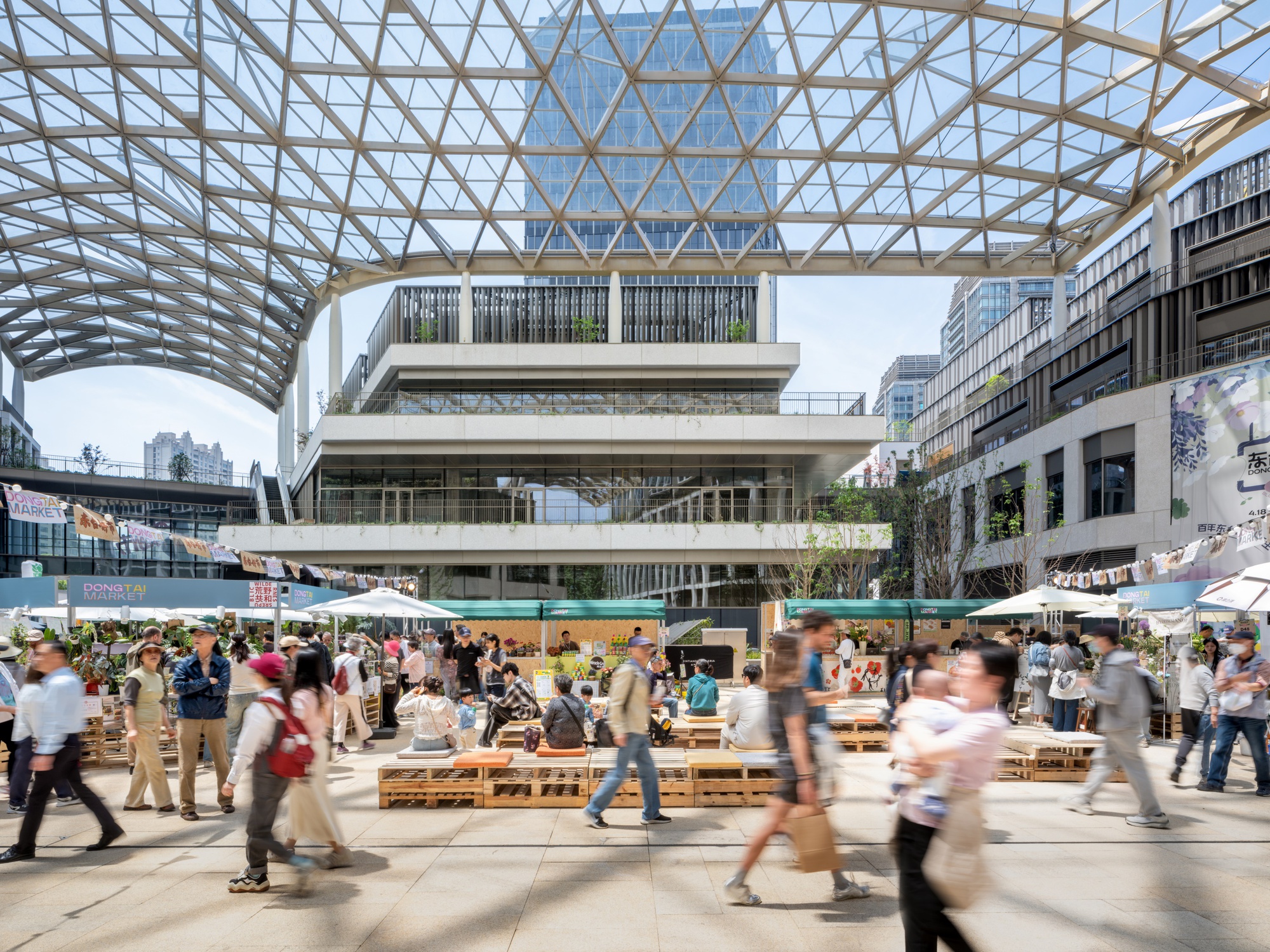 Covered plaza with diagonal lattice canopy sheltering crowds at wooden bench seating below terraced retail facades