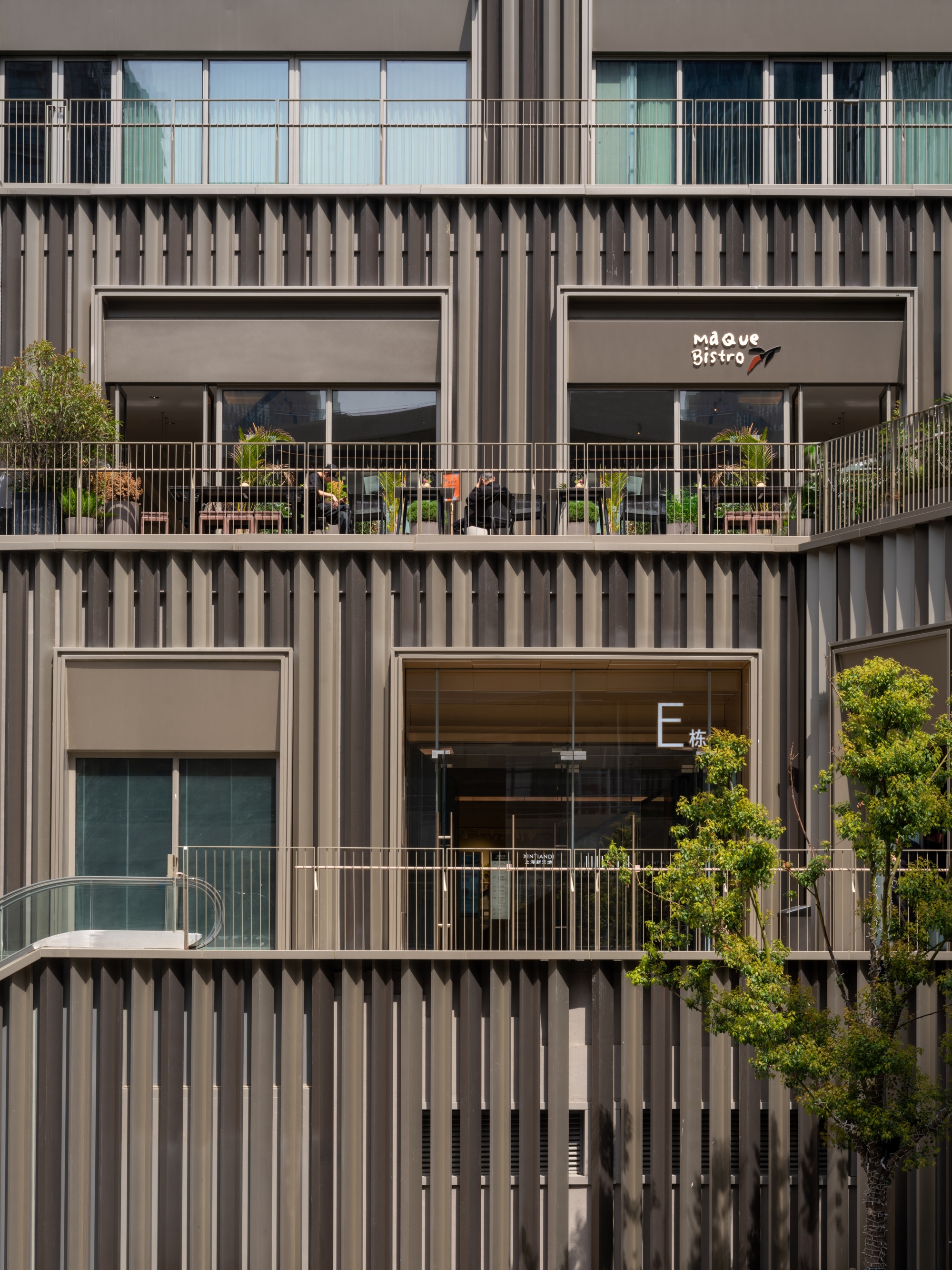 Stacked facade with vertical metal fins, recessed balconies, and potted trees on terrace railings