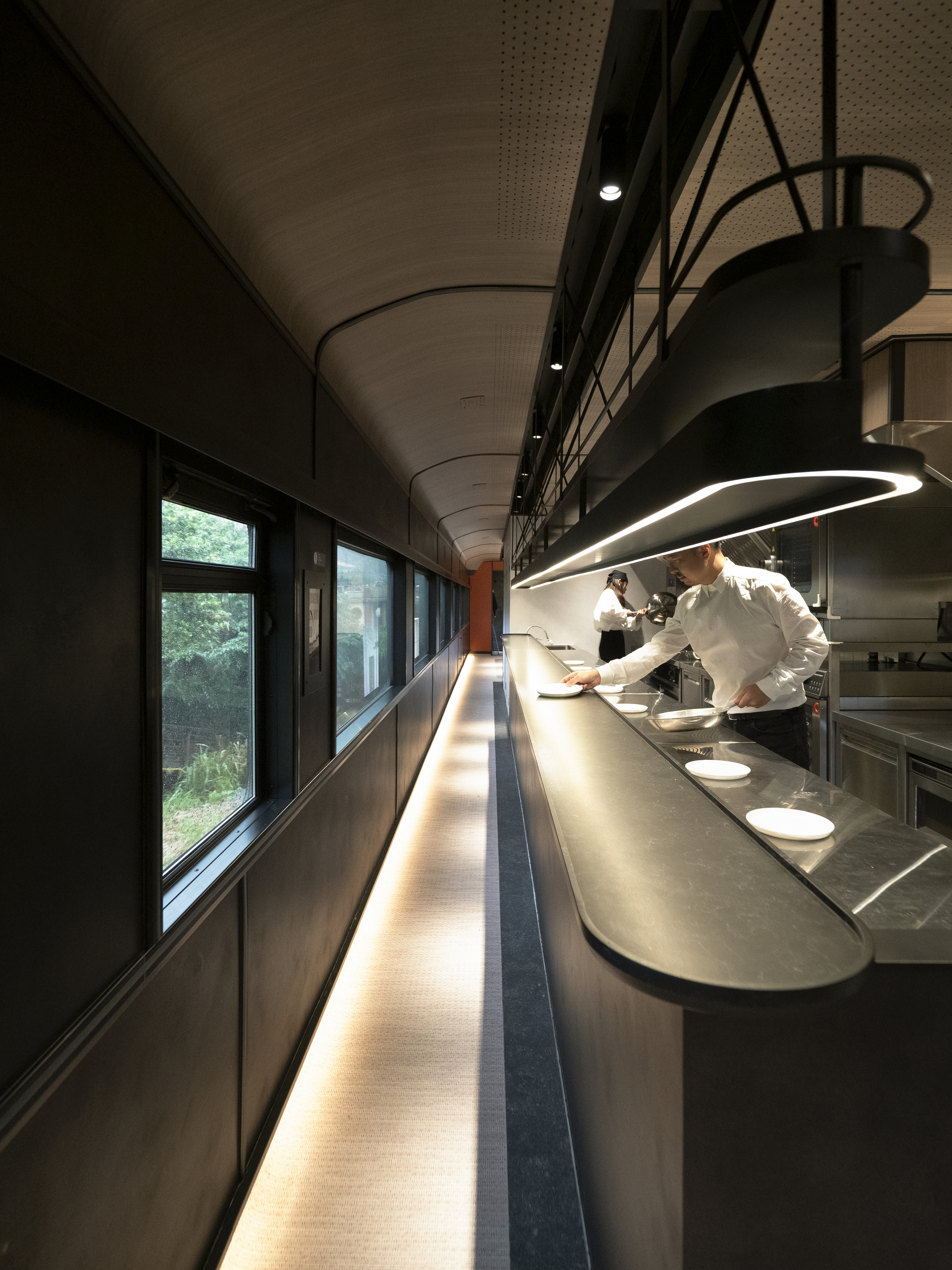 Galley workspace with continuous illuminated counter and chef plating under a curved ring pendant light