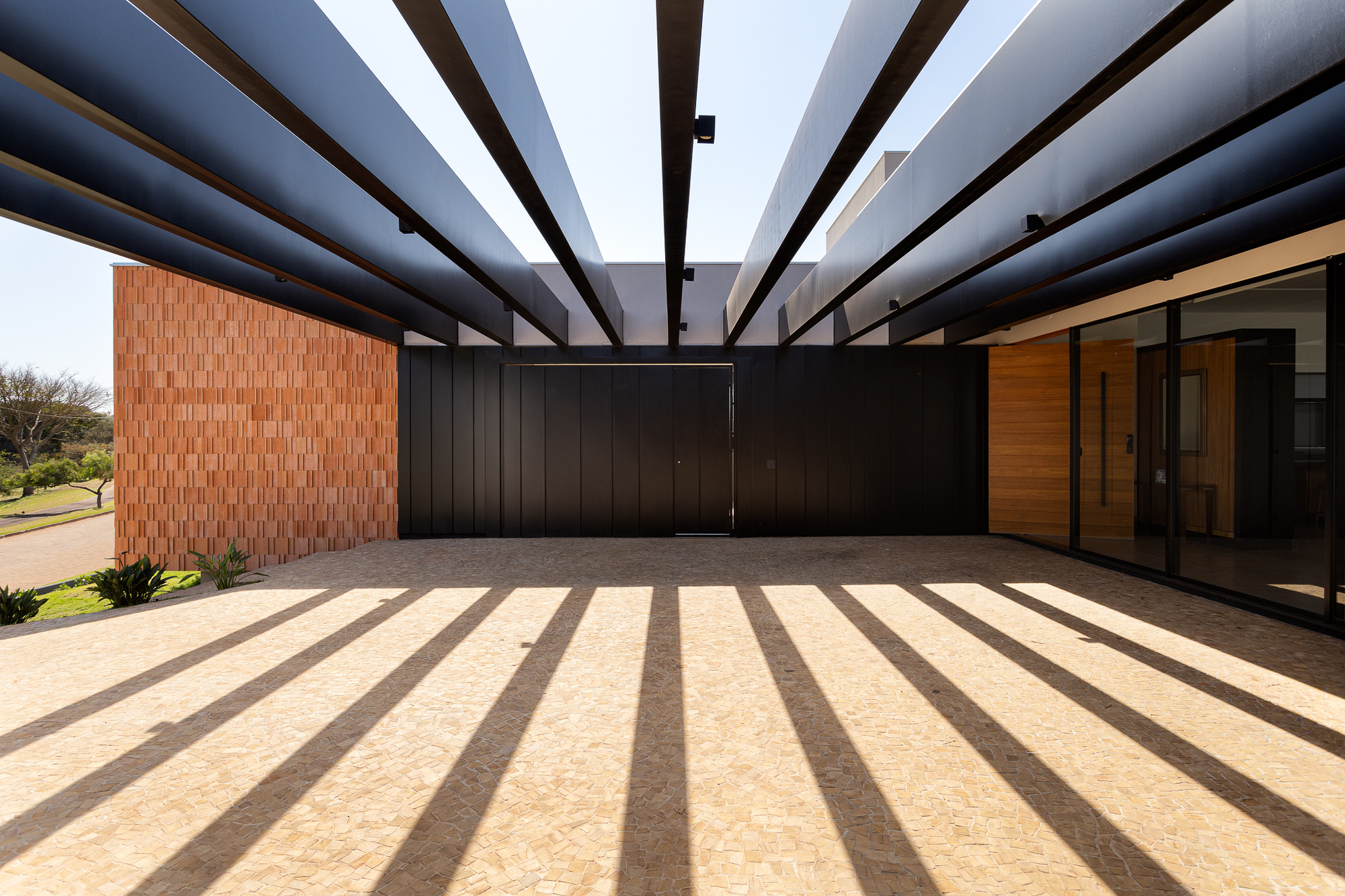 Covered entry courtyard with slatted overhead structure casting striped shadows across stone paving at midday