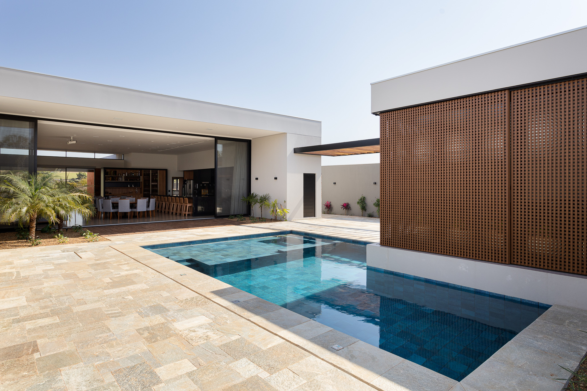 Courtyard pool with perforated timber screen wall, white stucco volumes, and sandstone paving under a clear sky