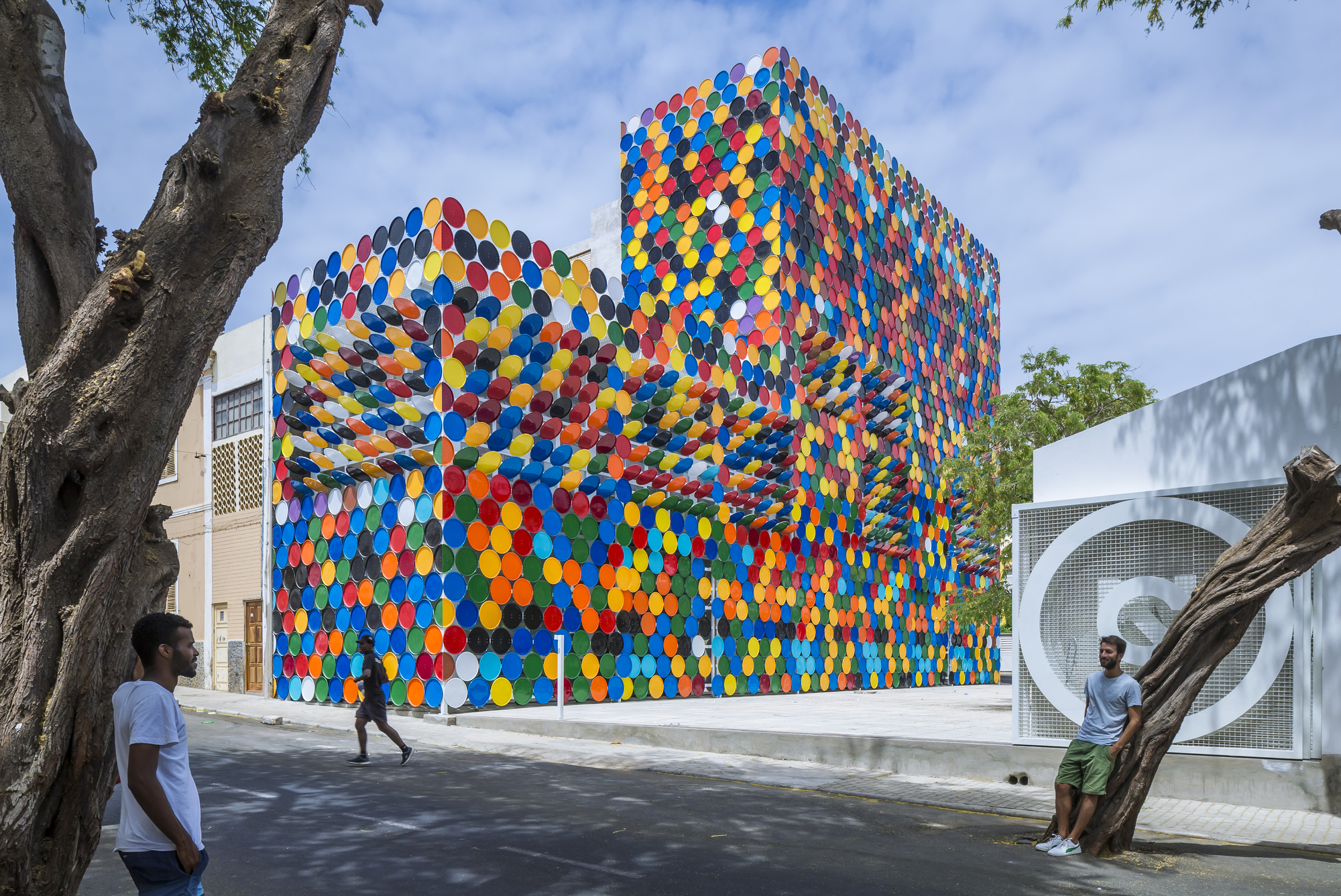 Street view of the facade clad in multicoloured circular discs beneath a tree and blue sky