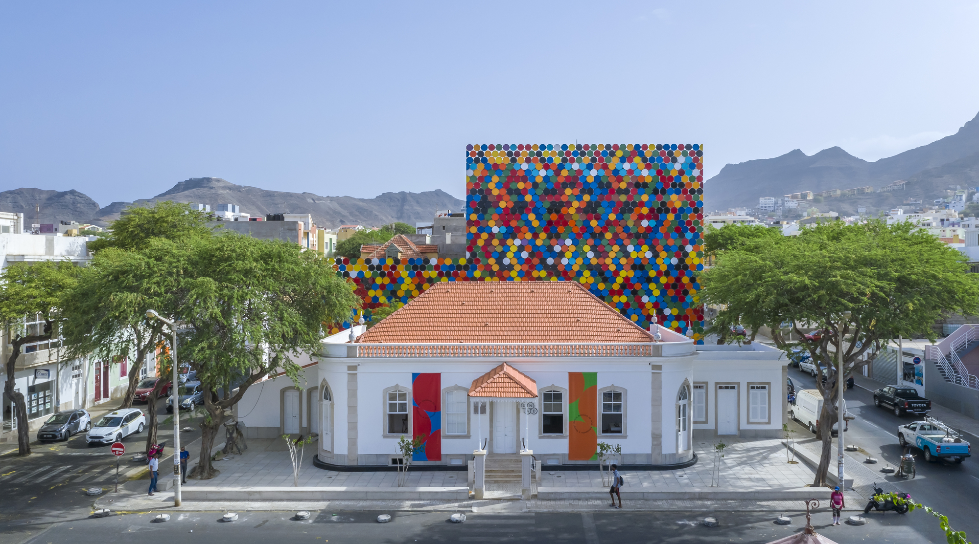 Aerial view showing the polychromatic disc facade rising behind a terracotta-roofed white building and green trees