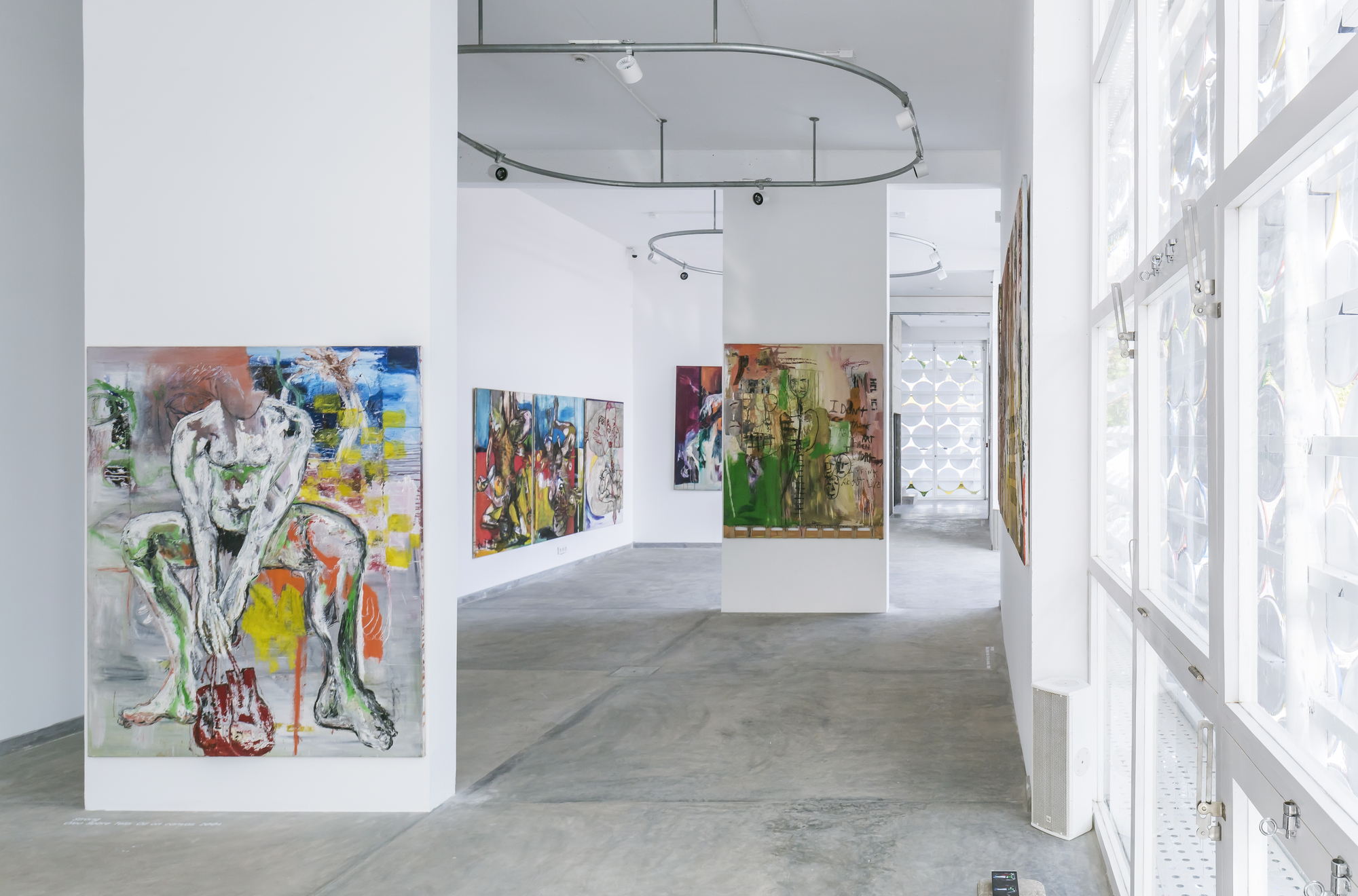 Gallery space with curved track lighting and paintings displayed along white walls beside the translucent screen facade