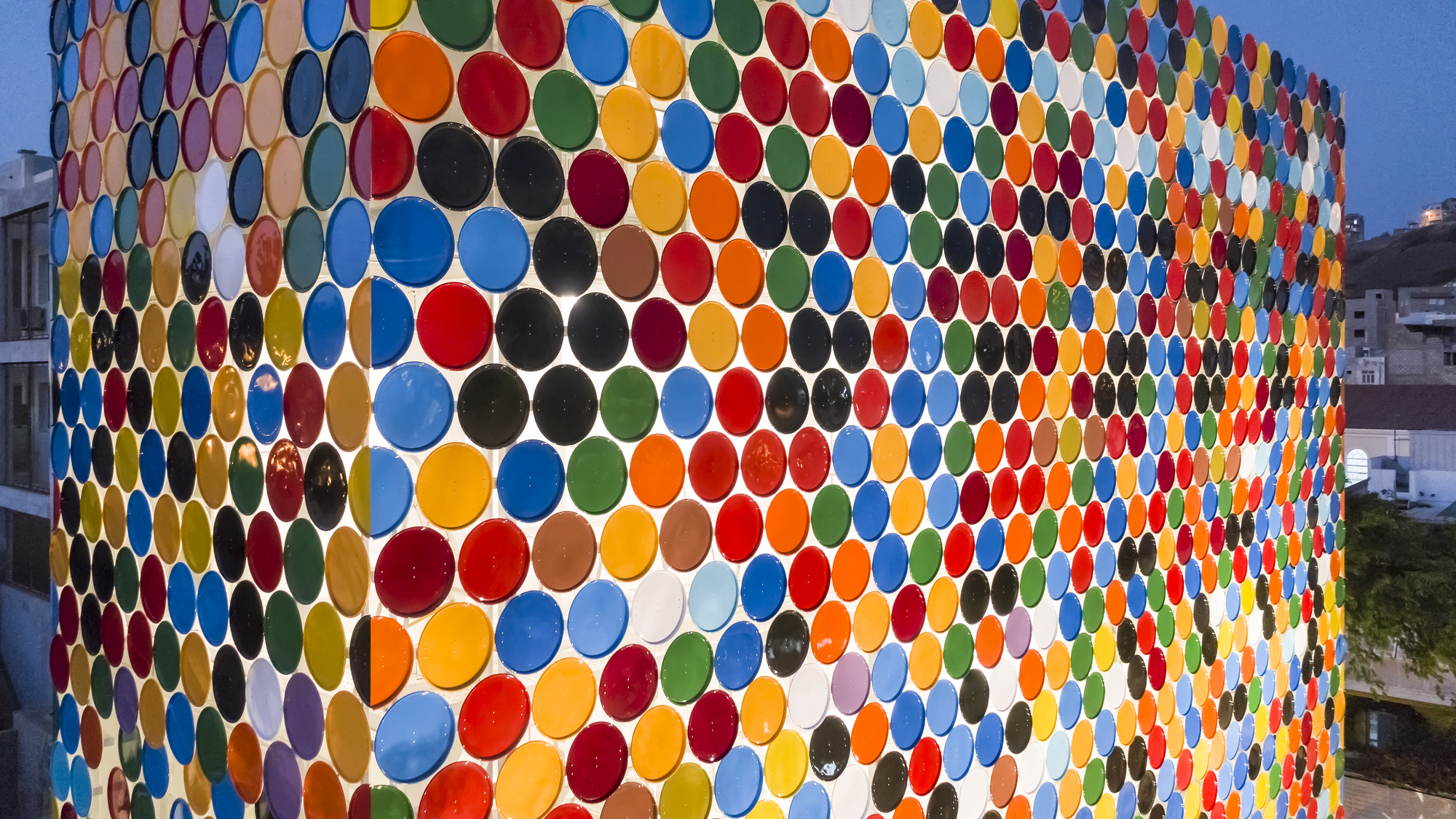 Facade clad in multicolored circular ceramic plates arranged in a honeycomb pattern at dusk