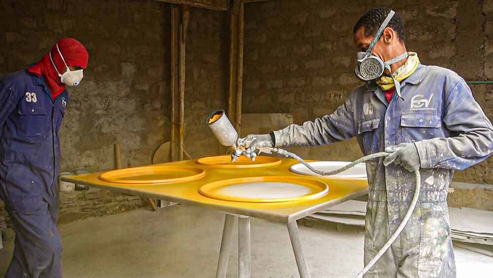 Worker spray-painting yellow finish onto panel with circular cutouts in a construction workshop